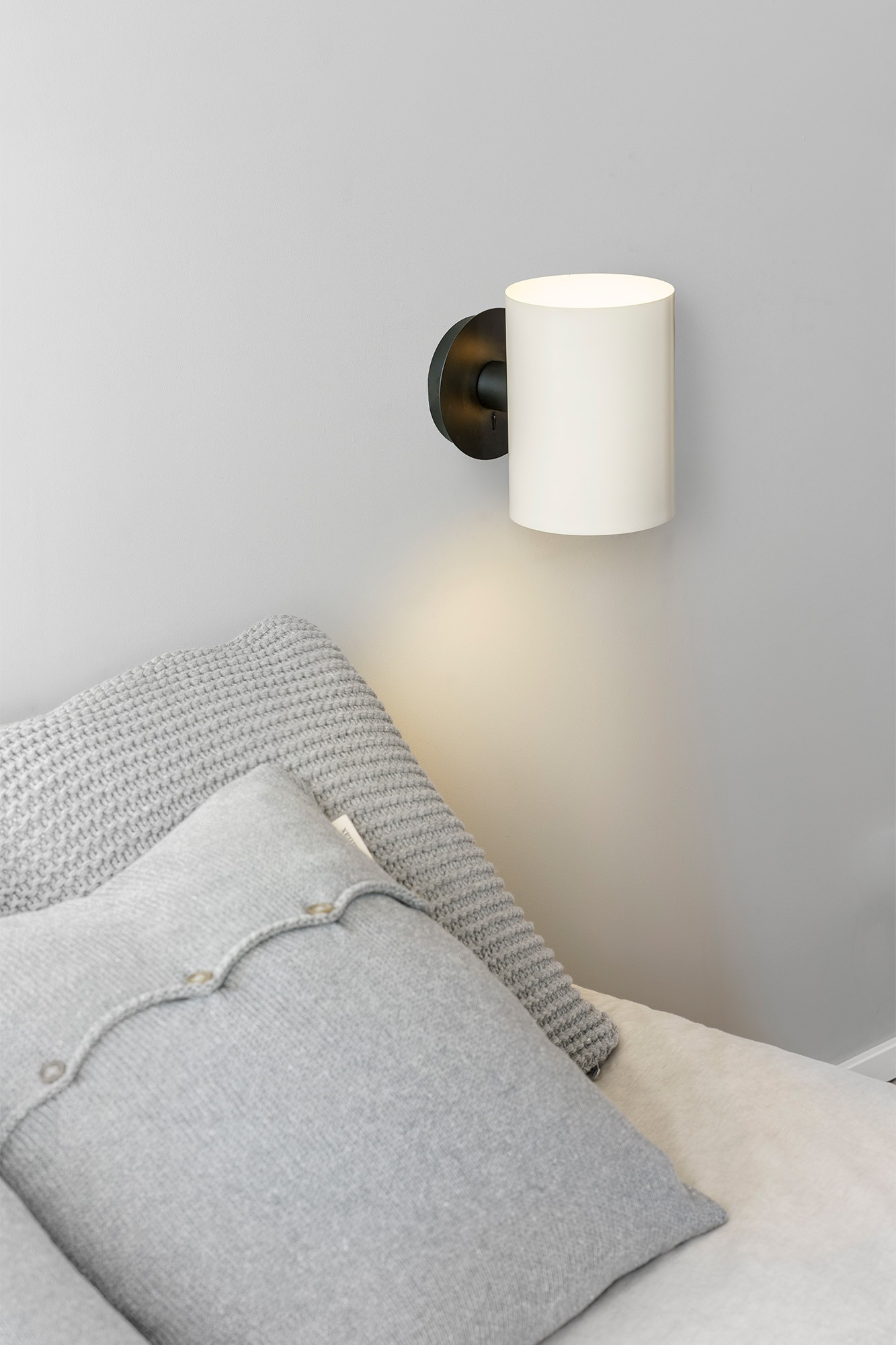 Lampada da parete cilindrica bianca sopra un cuscino grigio su un letto. Parete grigia sullo sfondo.