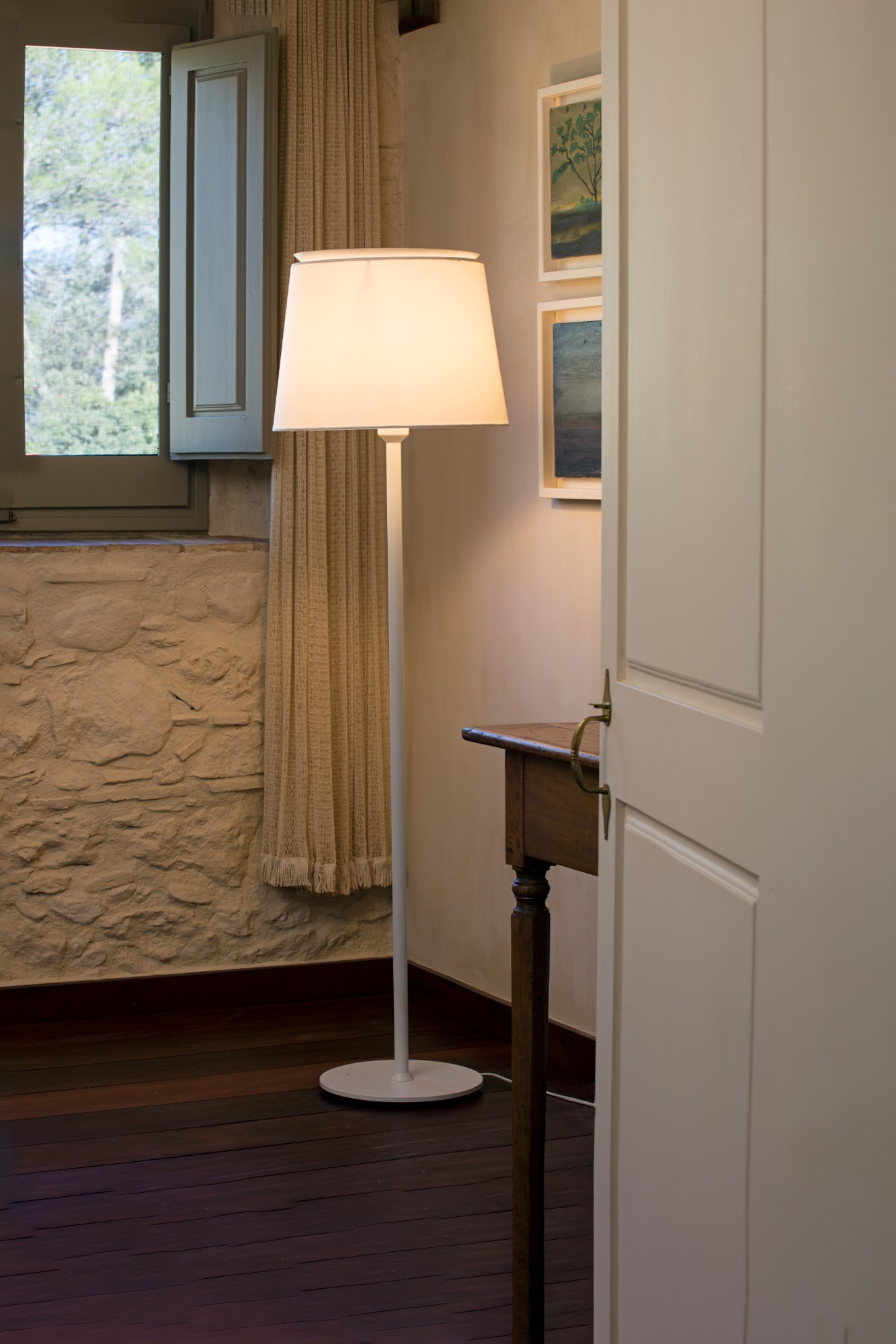 Una stanza con lampada, finestra e mobili in legno. La lampada ha un paralume bianco. La finestra ha delle persiane.
