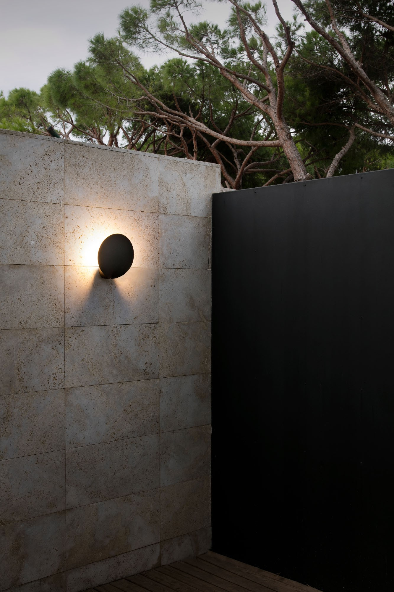 Una lampada da parete esterna rotonda e nera illumina un muro di pietra, completata da una recinzione scura e alberi.