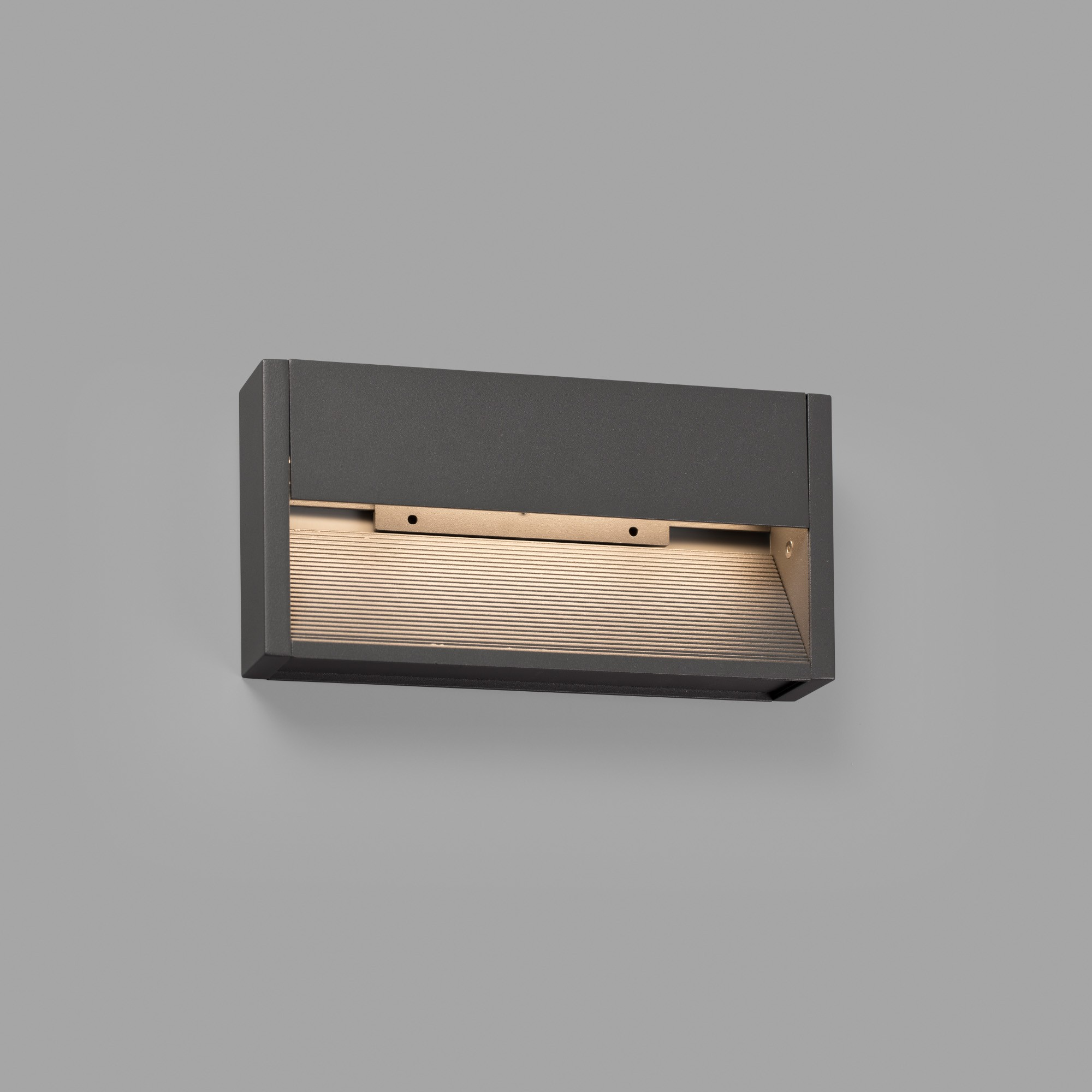 Un apparecchio di illuminazione rettangolare grigio scuro per esterni è montato su una parete grigio chiaro, con un'apertura illuminata.