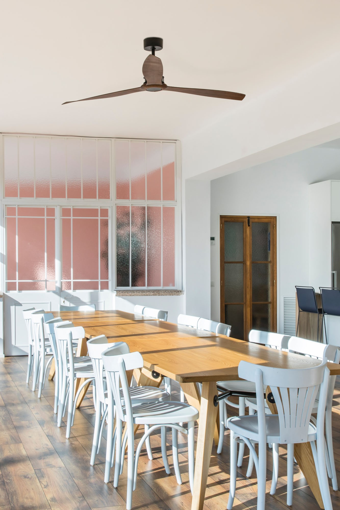 Una sala da pranzo con ventilatore a soffitto, grandi tavoli in legno, sedie bianche e una parete rosa.