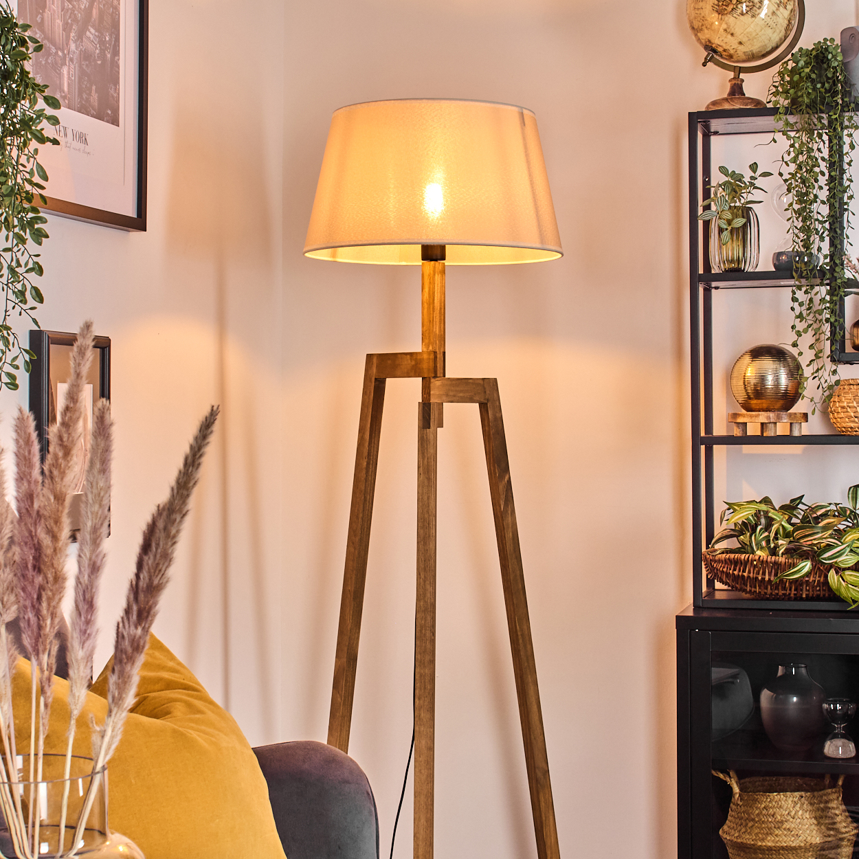 Eine hölzerne Stativlampe mit beleuchtetem Lampenschirm, in einem Raum mit Regal und Pflanzen.