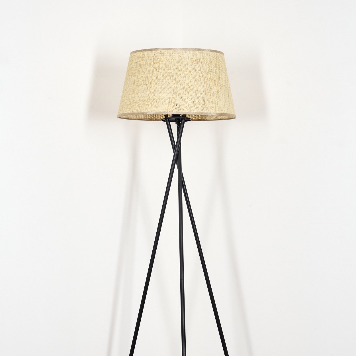 Eine schwarzbeinige Stehlampe mit beigem, zylindrischem Lampenschirm vor einer weißen Wand.