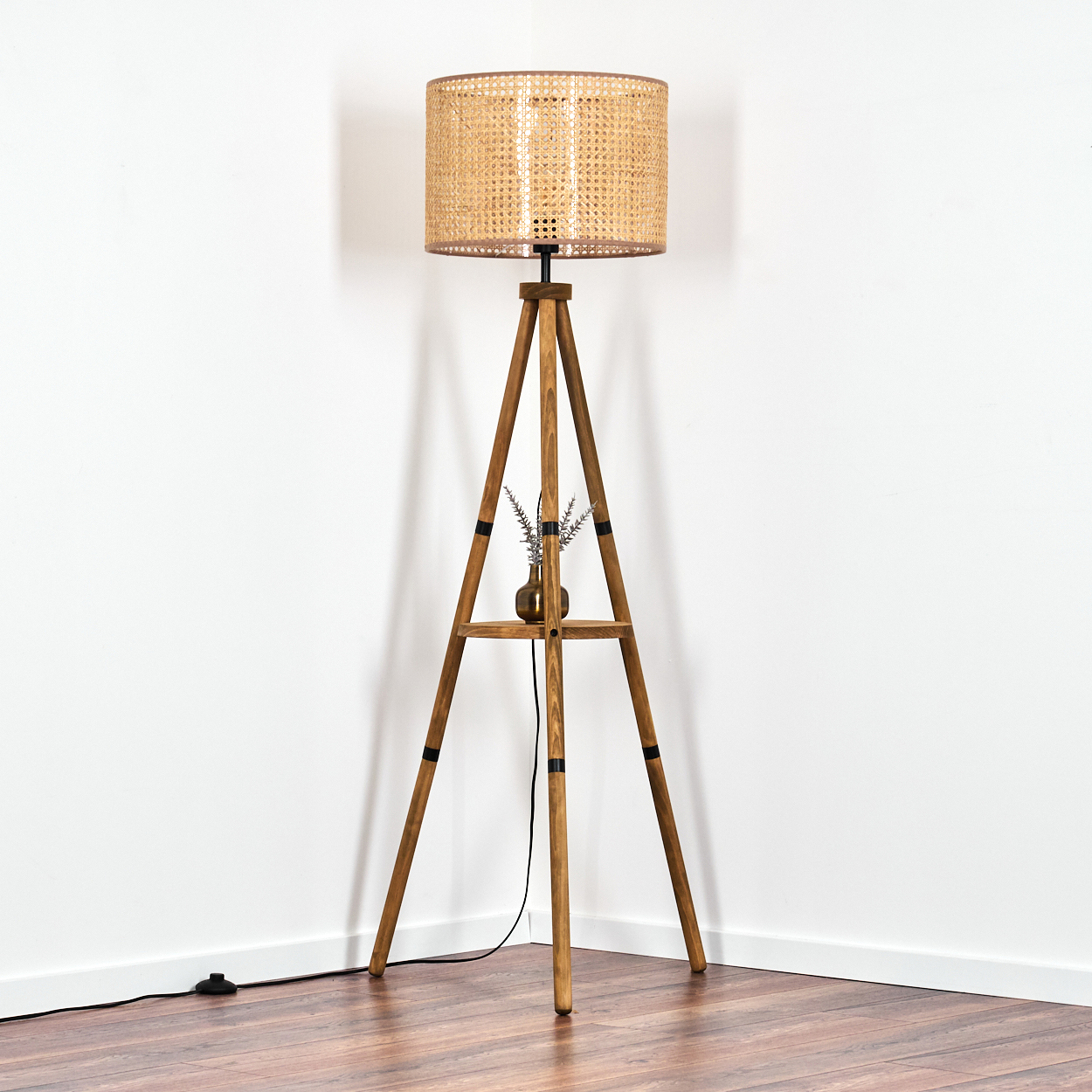 Eine hölzerne Stativ-Stehlampe mit einem Korblampenschirm und einer Blumenvase auf einem Regal.