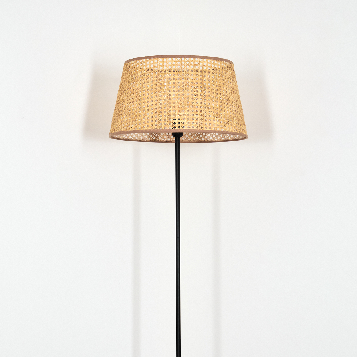 Stehlampe mit gewebtem Lampenschirm. Rattan-Textur, schwarzes Gestell, weißer Wandhintergrund.