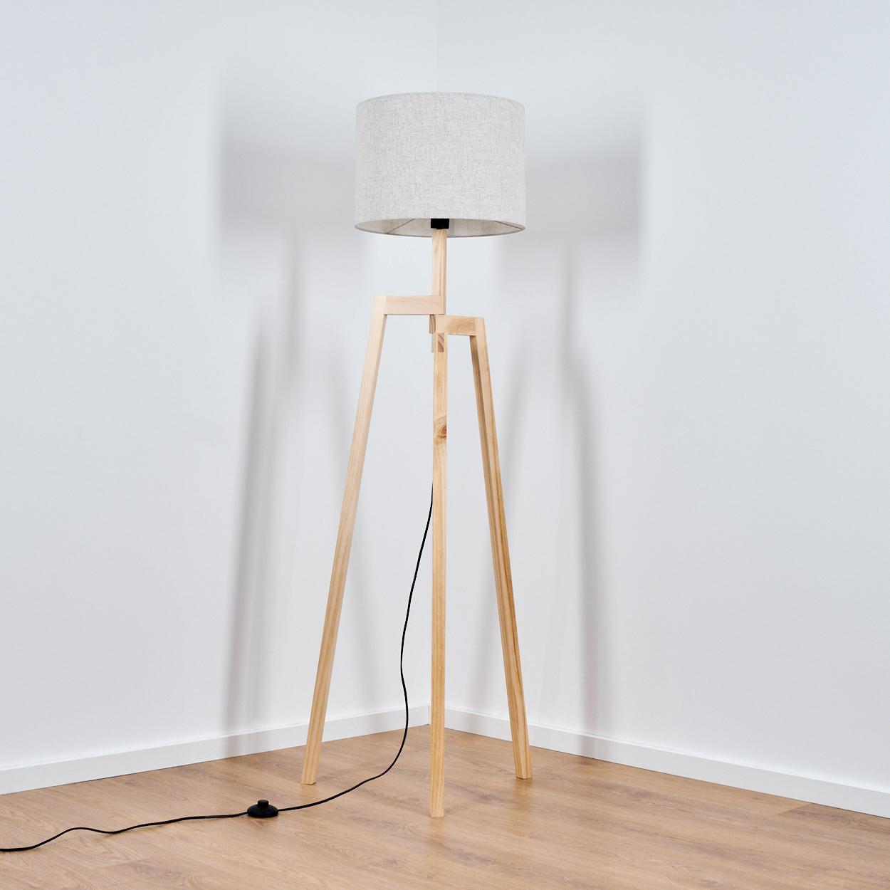 Dreibein-Stehlampe aus Holz mit grauem Lampenschirm. Die Lampe steht in einer Ecke nahe einer weißen Wand.