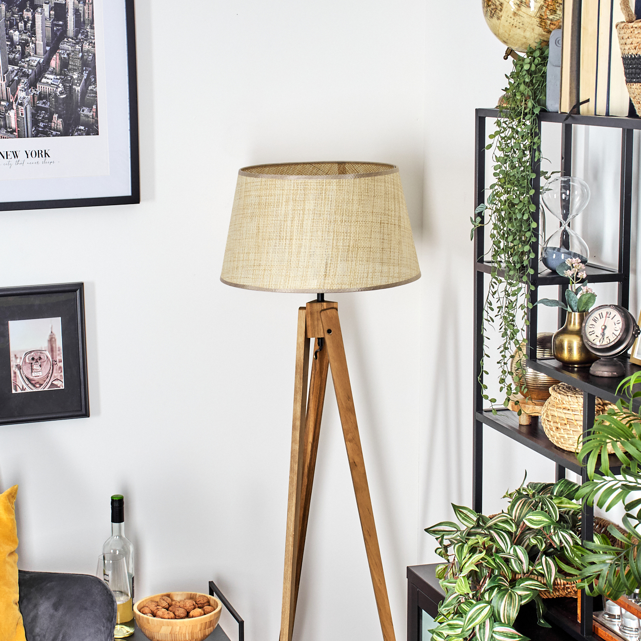 Dreibein-Stehlampe aus Holz mit beigefarbenem Lampenschirm, vor einer weißen Wand mit Dekoration.