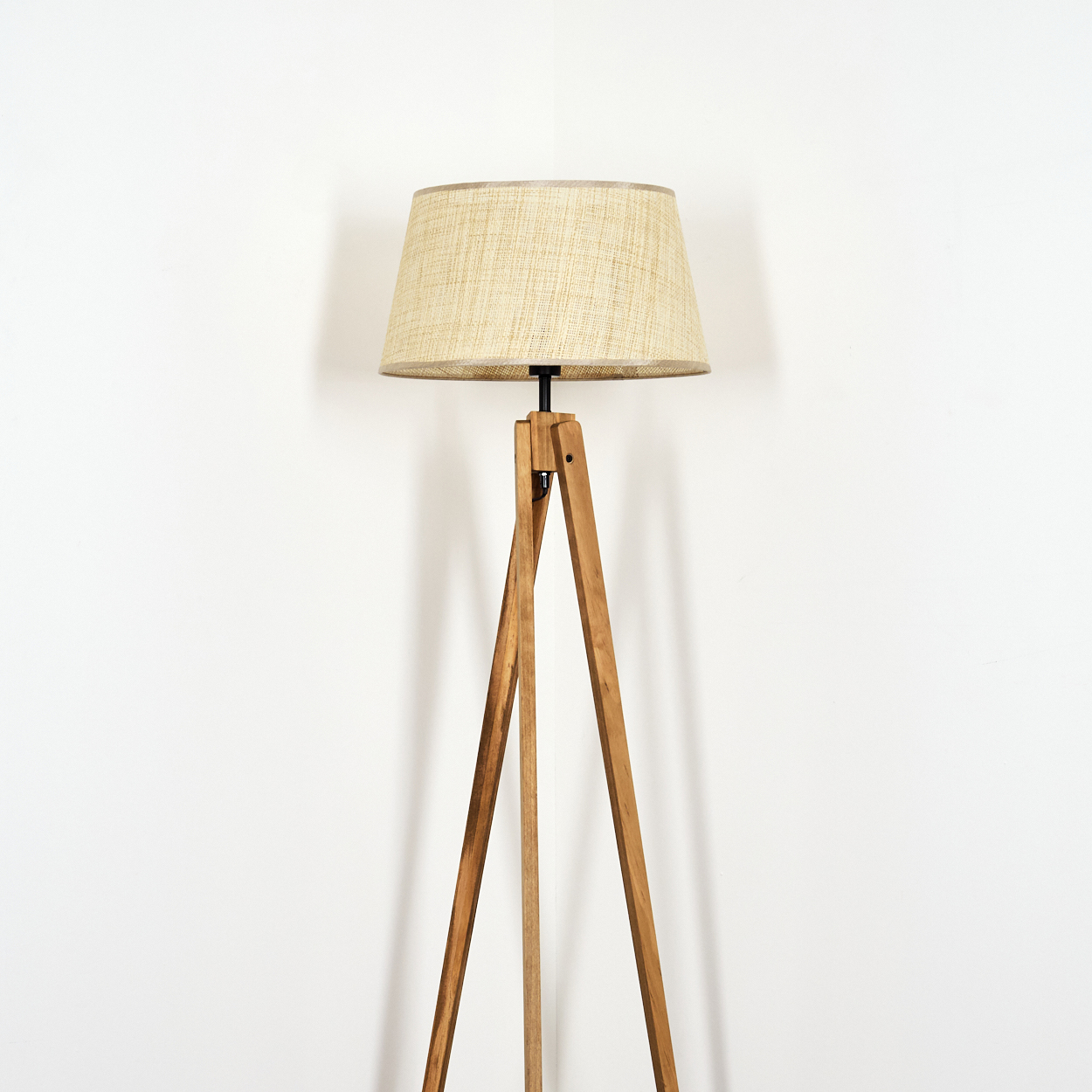 Holz-Dreibein-Stehlampe mit beigem, gewebtem Lampenschirm vor weißer Wand.