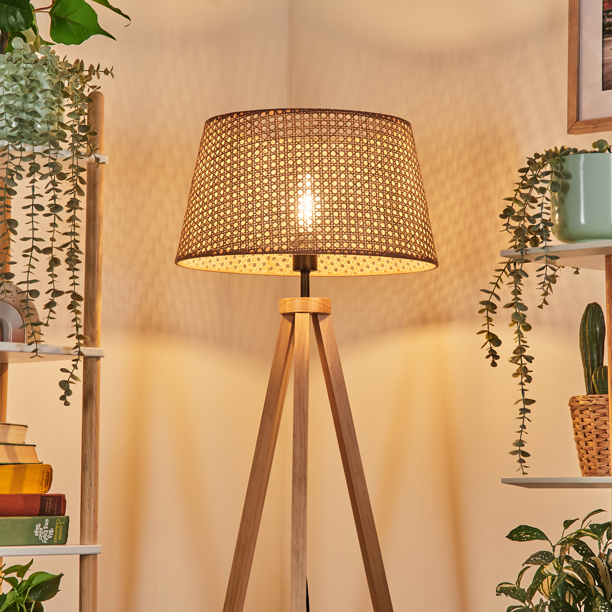Eine beleuchtete Stehlampe mit gewebtem Lampenschirm. Sie hat einen Holzstativfuß. Pflanzen sind um die Lampe sichtbar.
