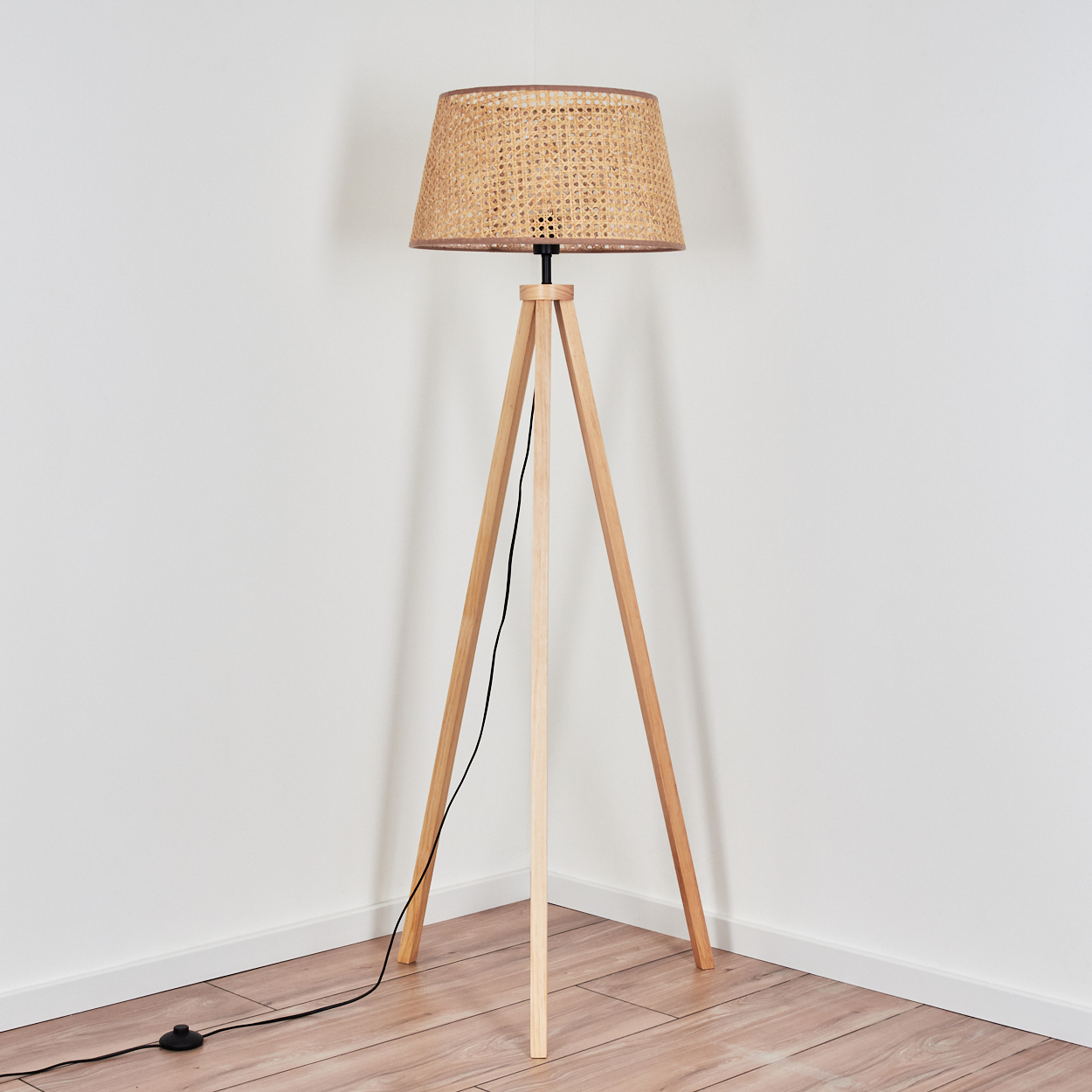 Eine Stehlampe in einer Ecke. Der Sockel ist aus Holz. Der Lampenschirm ist gewebt.