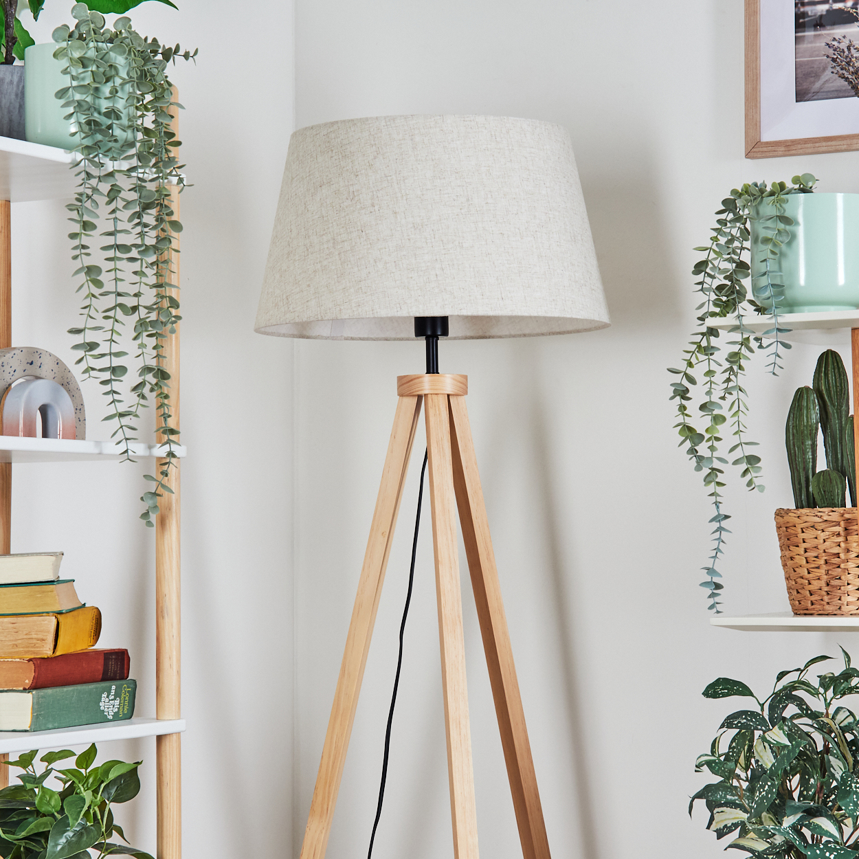 Eine Stativ-Stehlampe mit beigem Schirm in einem Raum mit Regalen und Pflanzen. Der Hintergrund ist weiß.