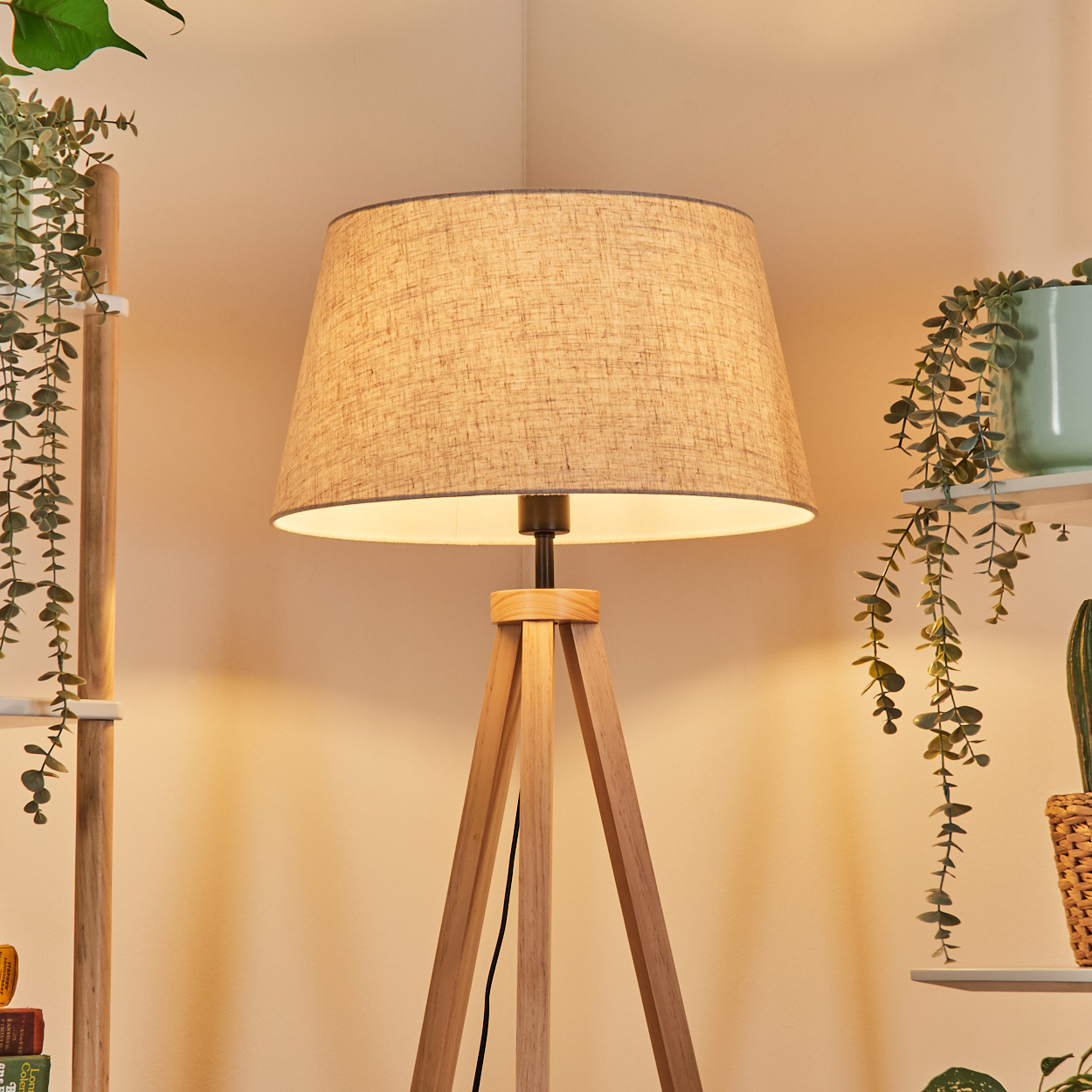 Stativ-Stehlampe in einer Zimmerecke. Pflanzen und Regale sichtbar. Helle Holzbeine, Stoffschirm, warmes Licht.