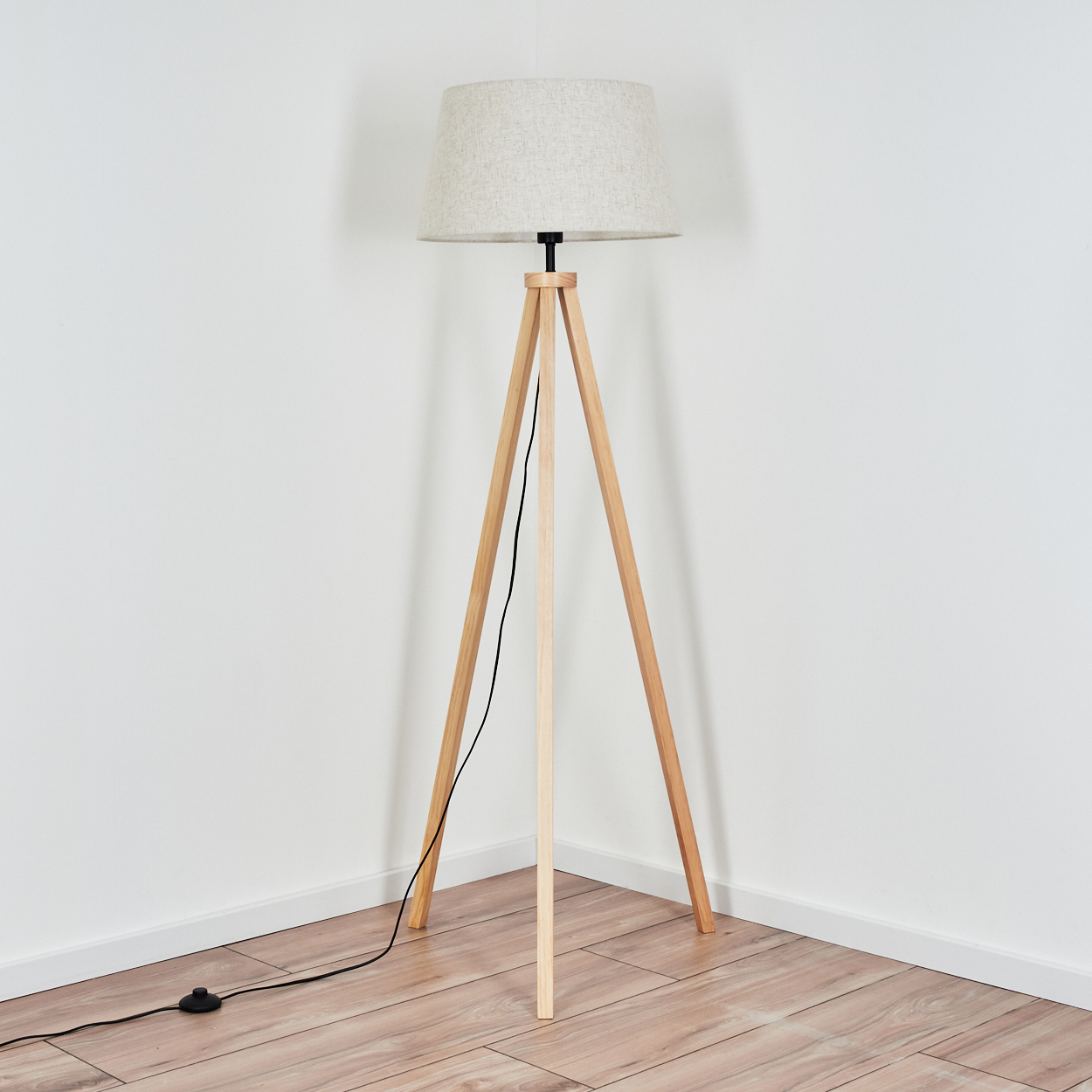 Eine hellfarbige Stehlampe aus Holz. Sie hat einen beigefarbenen Lampenschirm und steht in einer weißen Ecke.