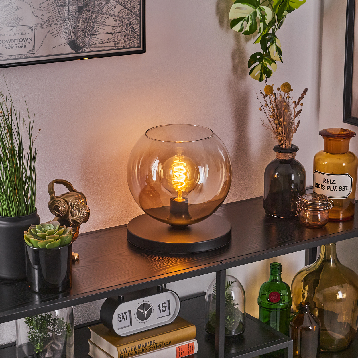 Eine Tischlampe mit bernsteinfarbenem Glasschirm, schwarzem Sockel, Glühbirne, auf Regal mit Deko.