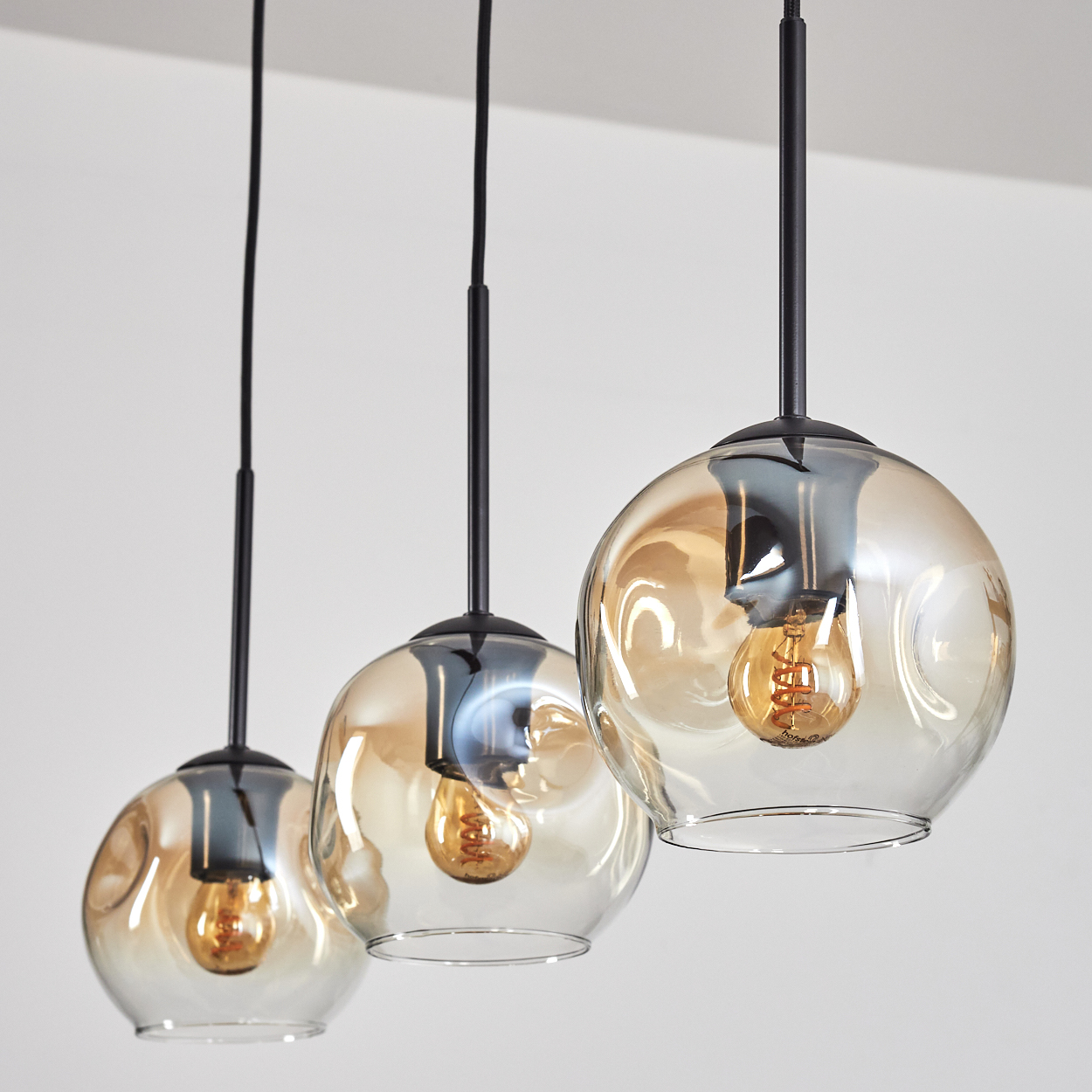 Drei Glas-Pendelleuchten mit bernsteinfarbenen Glühbirnen, an schwarzen Kabeln, vor weißem Hintergrund.