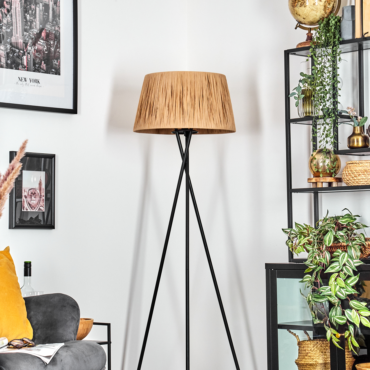 Eine Stehlampe mit beigefarbenem Lampenschirm und schwarzen Beinen steht in einem Raum mit Möbeln und Pflanzen.