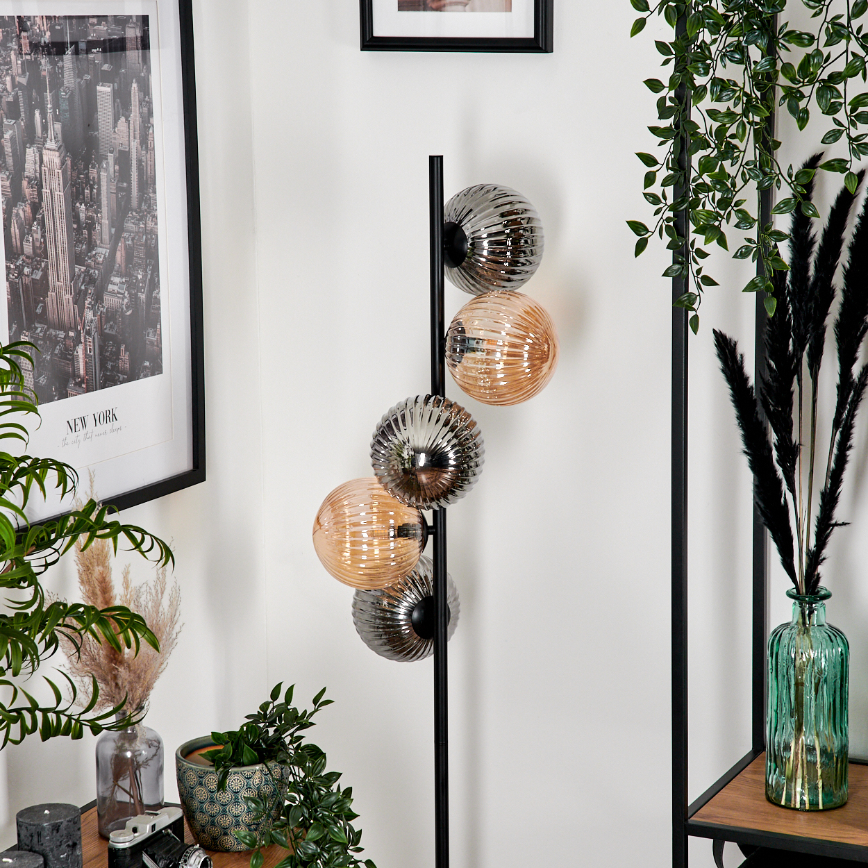 Eine Stehlampe mit vier Kugelschirmen, abwechselnd silber und bernsteinfarben, steht vor einer weißen Wand. Pflanzen und ein New York-Druck sind in der Nähe.