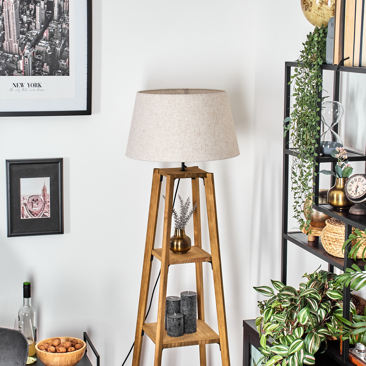Holz-Stehlampe mit zwei Regalen. Beiger Lampenschirm, kleine Vase und Kerzen auf den Regalen. Weiße Wand im Hintergrund.