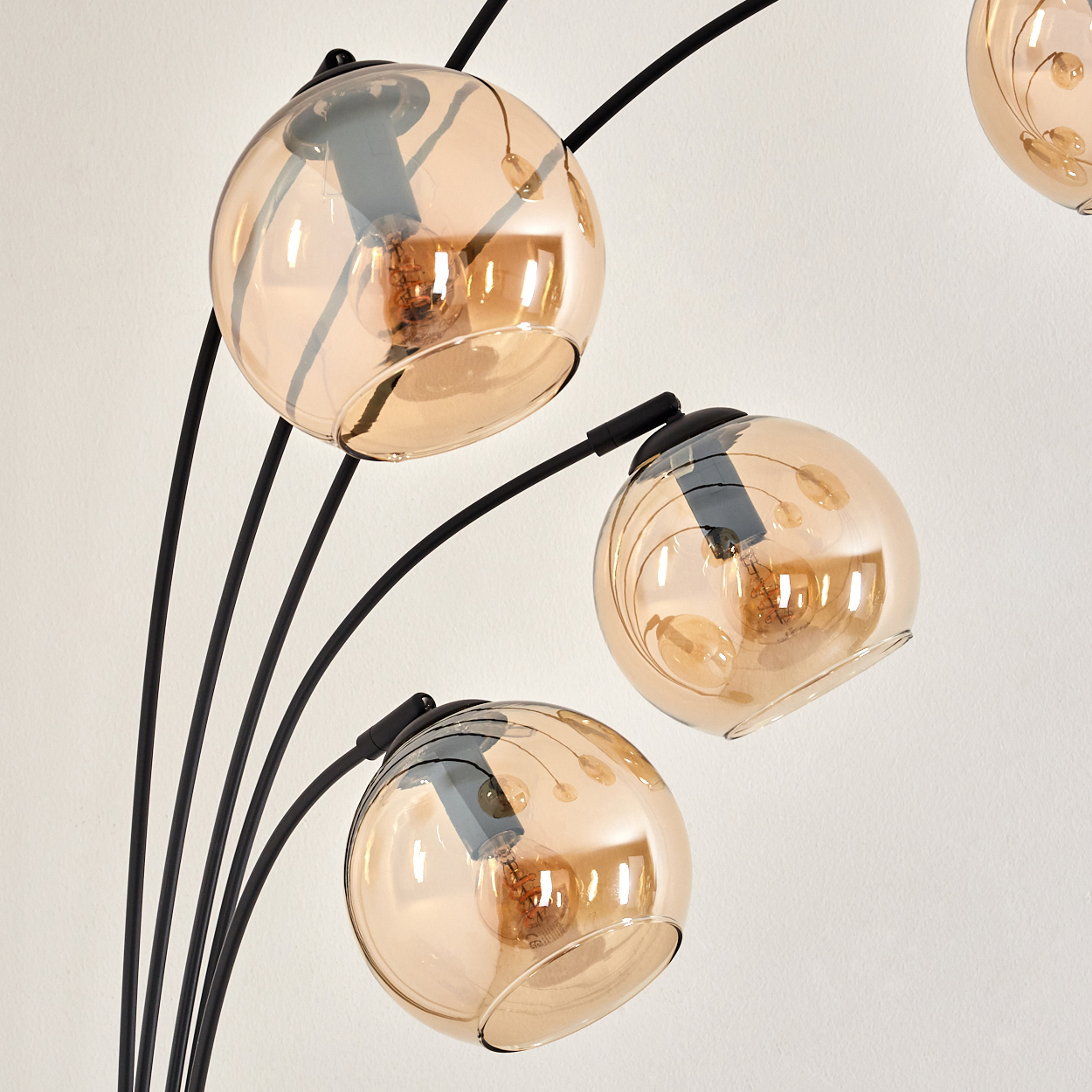 Nahaufnahme einer schwarzen Stehlampe mit fünf bernsteinfarbenen Glaskugeln, beleuchtet, vor weißer Wand.