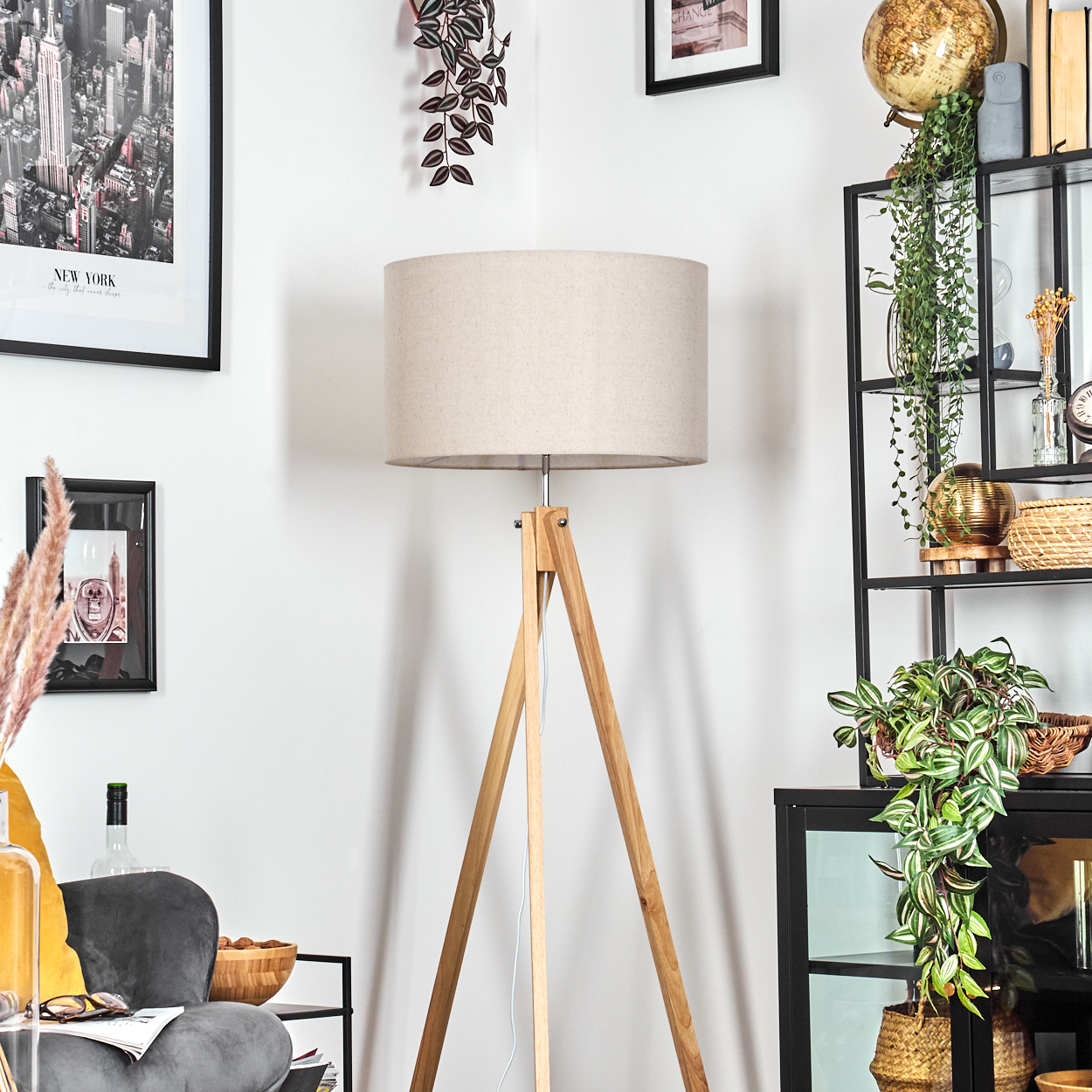Wohnzimmerszene mit Holz-Stativlampe, beigem Schirm und verschiedenen Dekorationsartikeln.