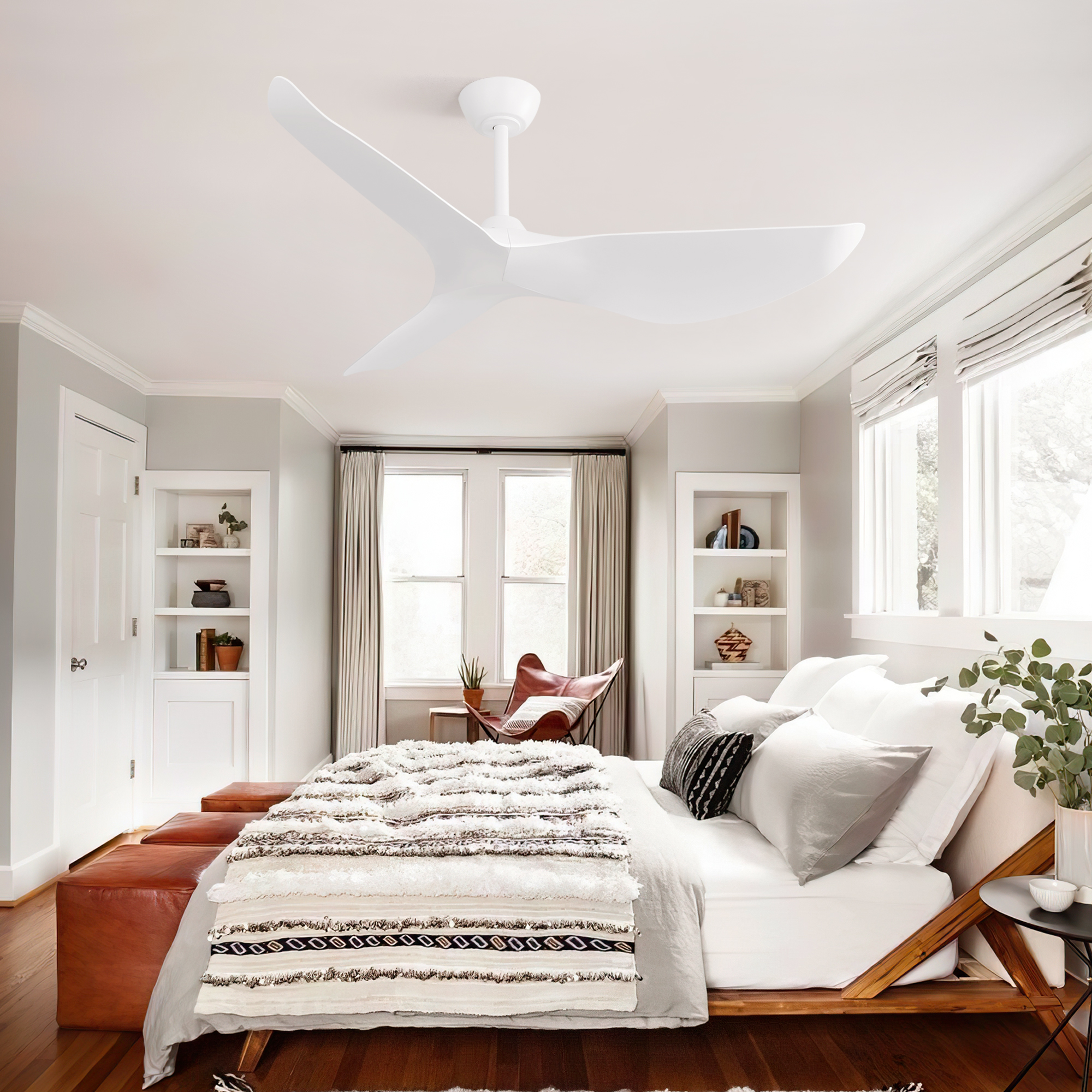 Ein Schlafzimmer mit weißem Deckenventilator, Bett und Fenstern. Die Einrichtung umfasst Pflanzen, neutrale Töne und eine Leder-Ottomane.