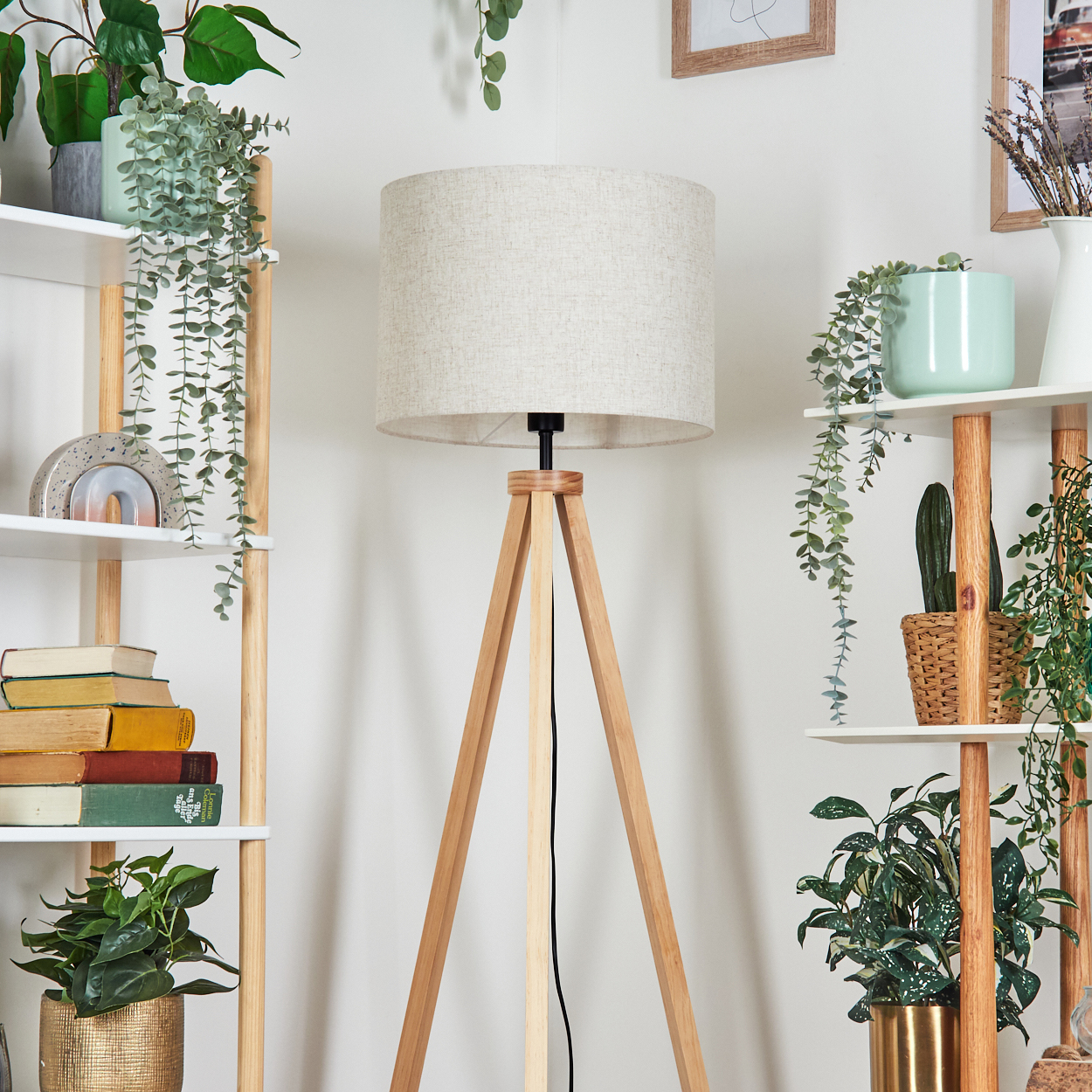 Eine hölzerne Stativ-Stehleuchte mit hellfarbigem Lampenschirm vor einer Wand mit Pflanzen.