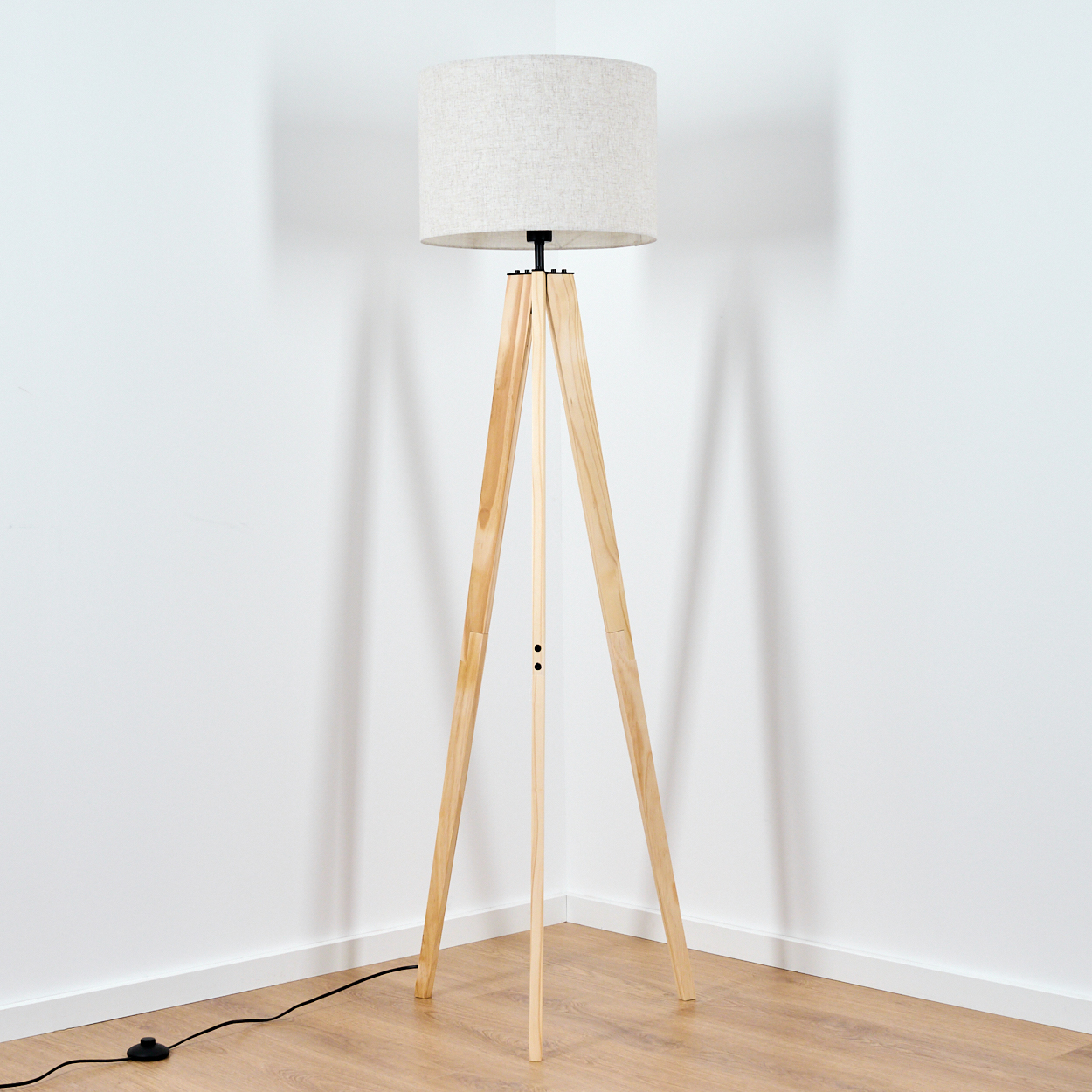 Eine Stehlampe mit einem Stativfuß aus Holz und einem hellen Lampenschirm aus Leinen, in einer Ecke.