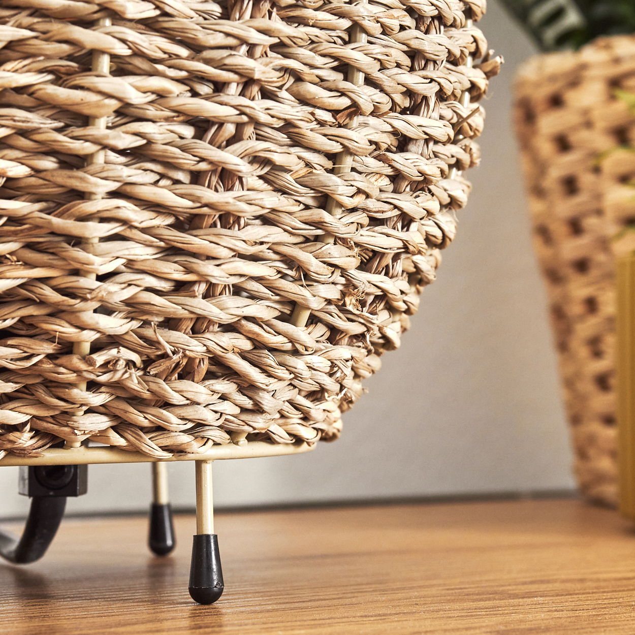 Gewebte beige Lampe auf Holzoberfläche, Beine und schwarzes Kabel zeigend, mit beigem Hintergrund.
