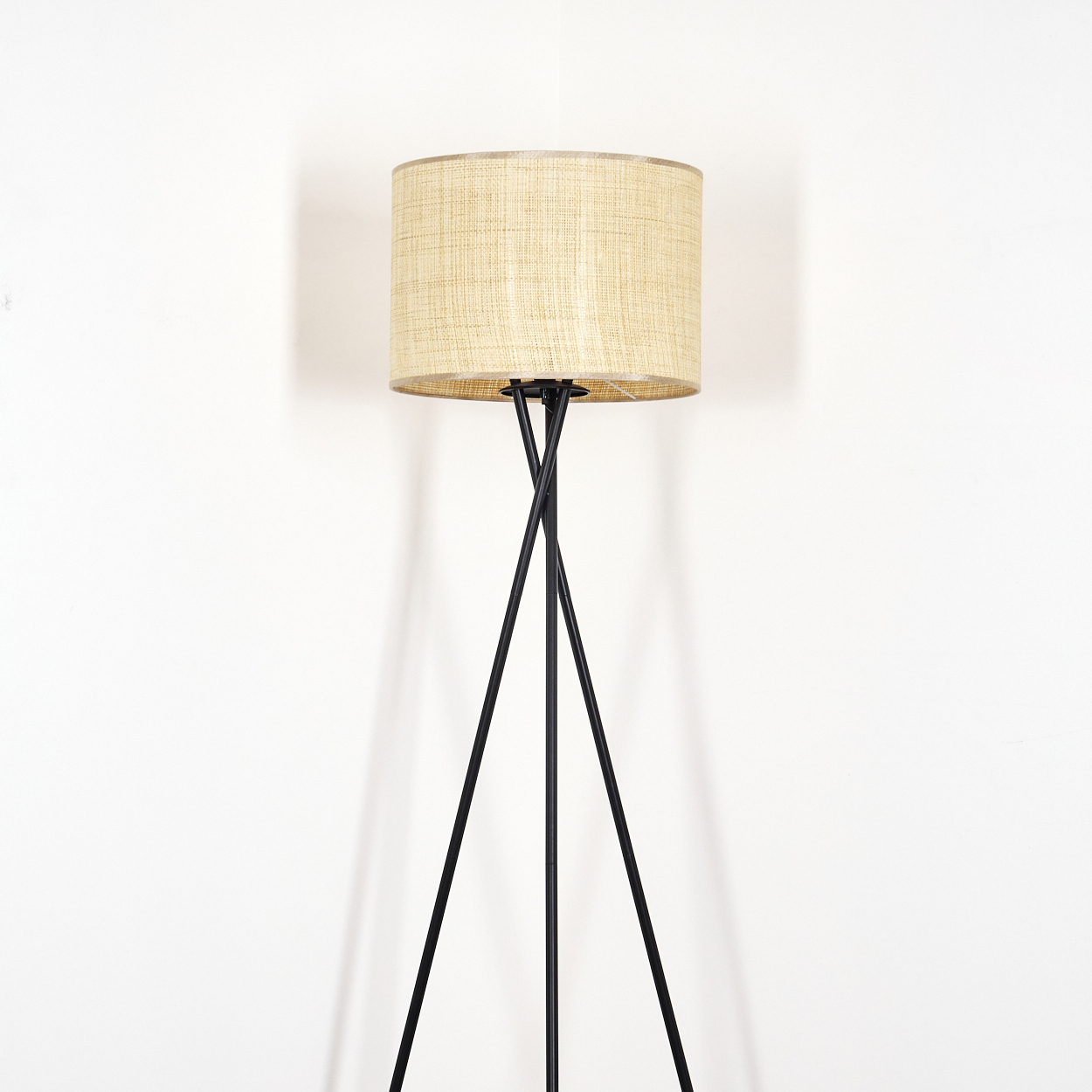 Eine Stehlampe mit beigem Lampenschirm auf drei schwarzen Beinen. Die Lampe steht vor einer weißen Wand.