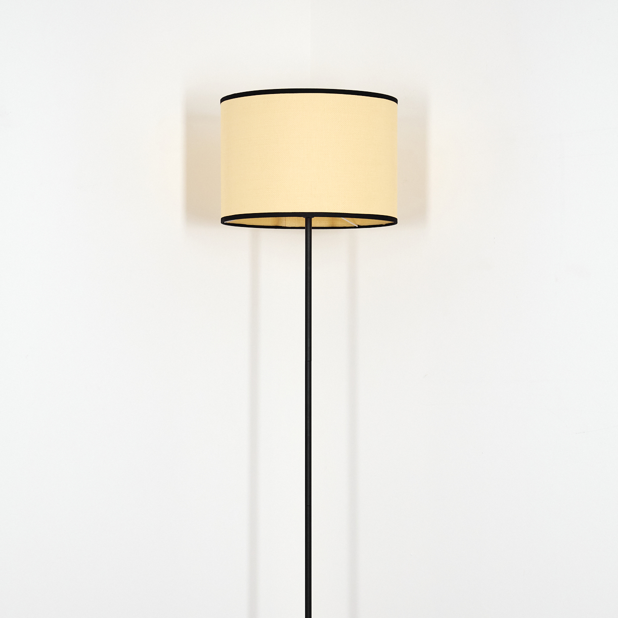Stehlampe mit beigem Lampenschirm mit schwarzem Rand, schwarzem Pol, vor weißer Wand.