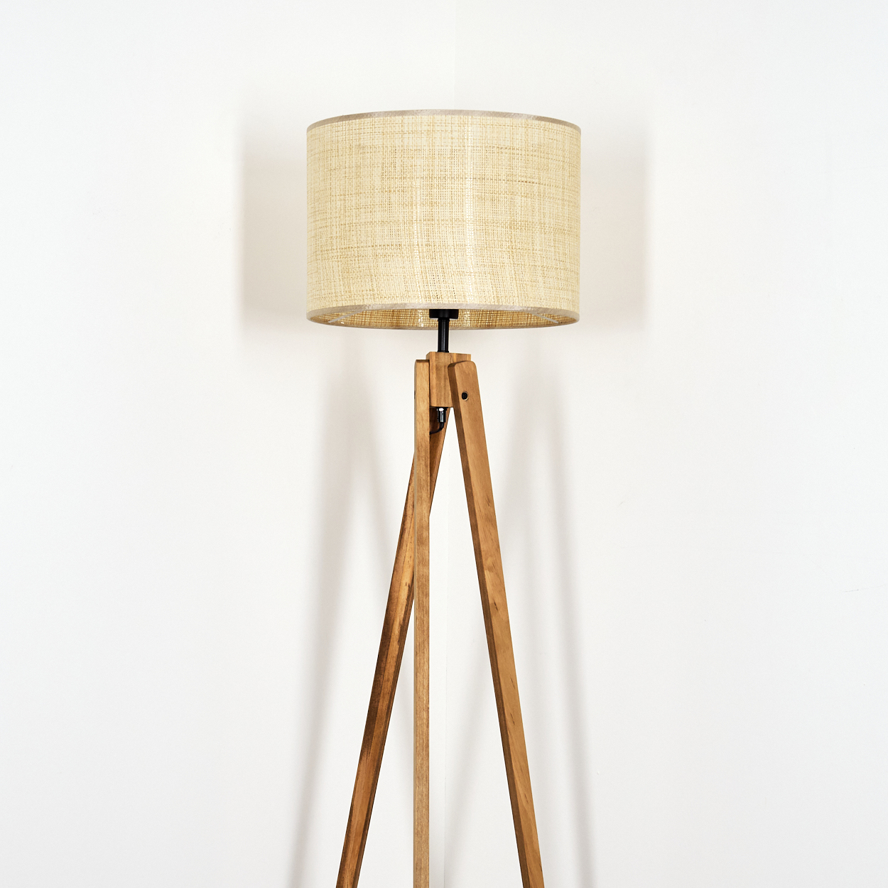Stehlampe mit beigem Schirm und Holz-Dreibein-Fuß vor weißer Wand.