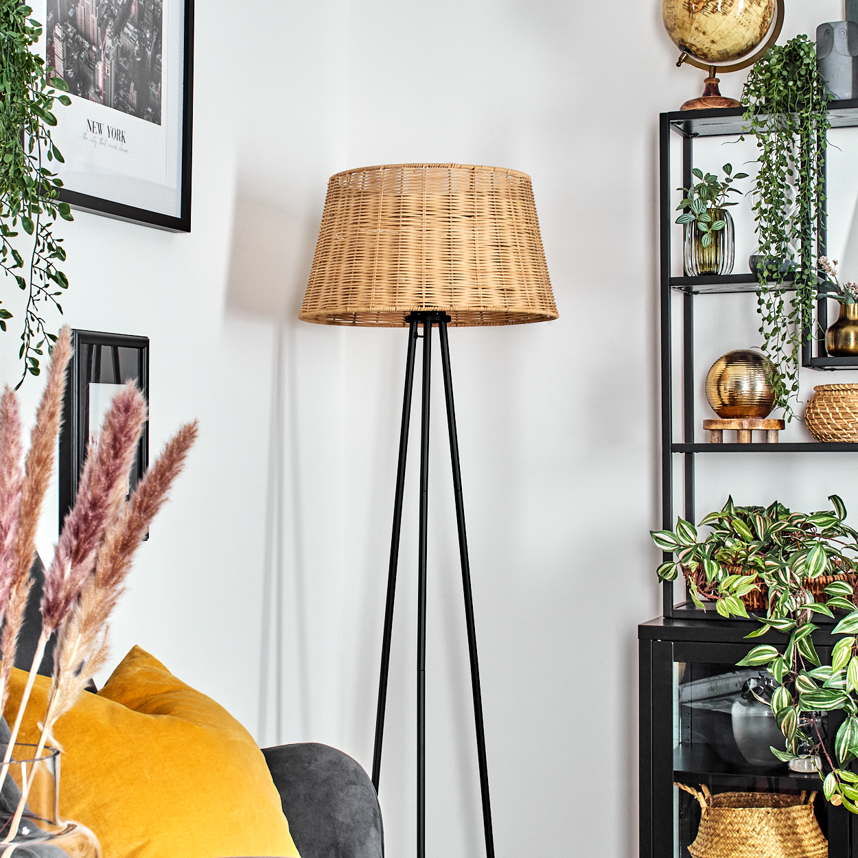 Eine Zimmerecke mit einer gewebten Lampe, einem schwarzen Regal mit Pflanzen, einem gelben Kissen und einem gerahmten Bild.
