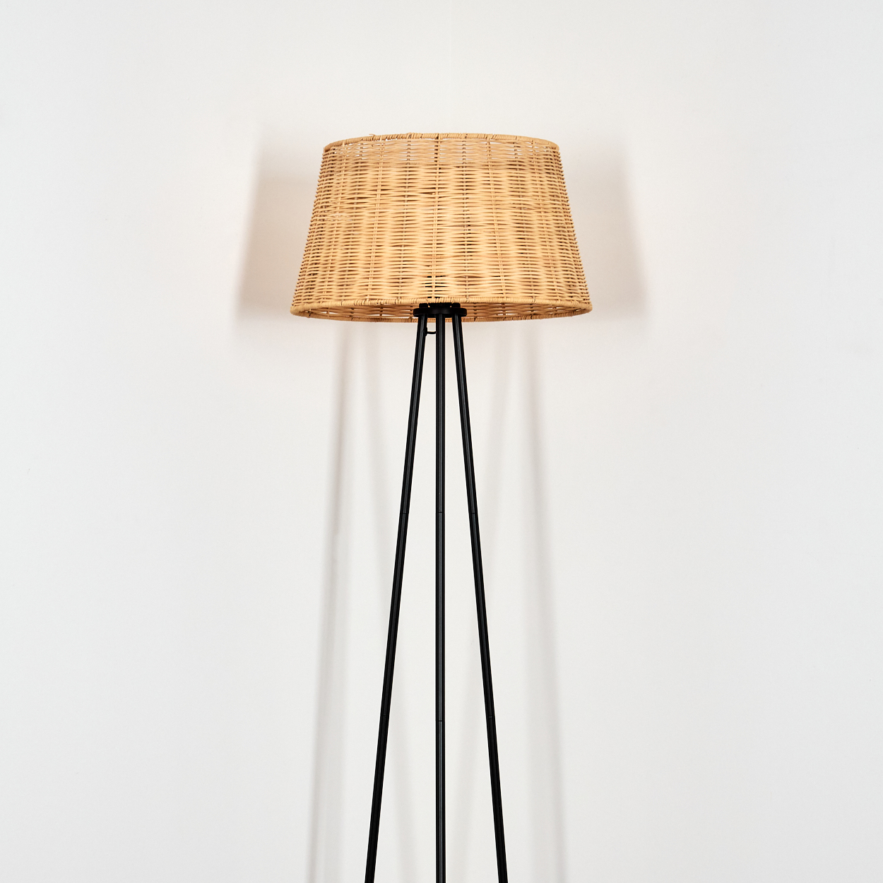 Eine hohe Stehlampe mit einem gewebten Lampenschirm auf schwarzen Beinen steht vor einer weißen Wand.
