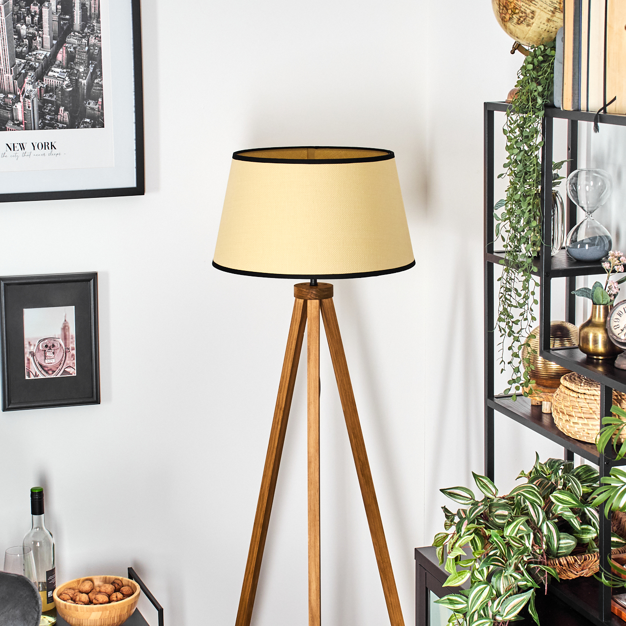 Eine hölzerne Dreibeinlampe mit beigem Schirm und schwarzem Besatz, in einem Wohnzimmer mit Deko.