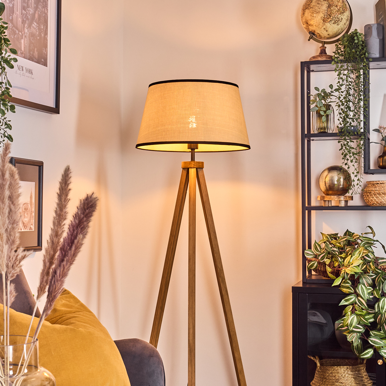 Eine hölzerne Dreibeinlampe mit beigem Schirm und schwarzem Besatz in einem Wohnzimmer.