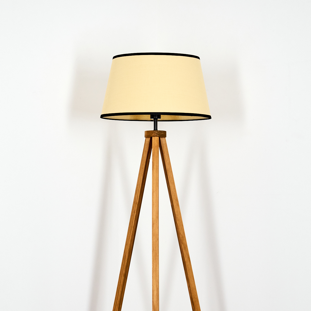 Eine hölzerne Dreibeinlampe mit beigem Schirm und schwarzem Besatz. Die Lampe steht vor einer weißen Wand.