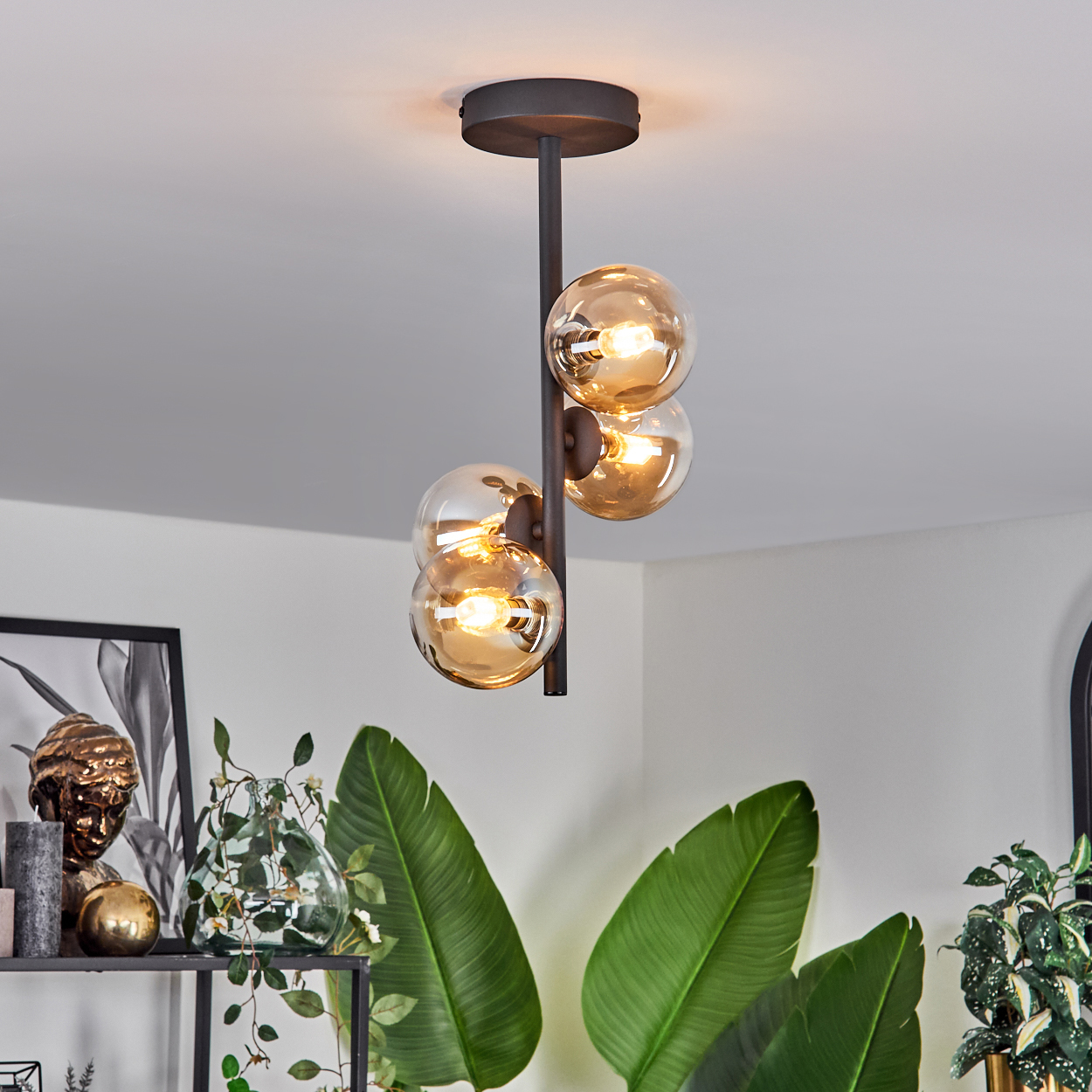 Deckenlampe mit braunem Rahmen, vier Glaskugeln und Hintergrund eines Zimmers mit Pflanzen.