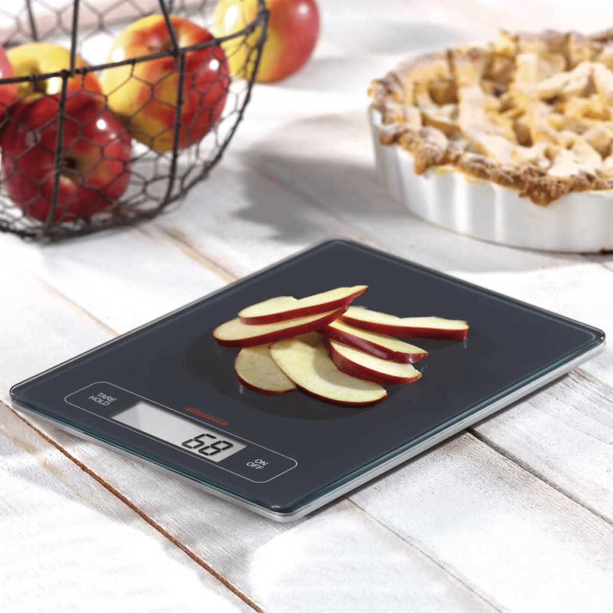 Waga kuchenna z plasterkami jabłka. Jabłka w koszyku i szarlotka w tle.