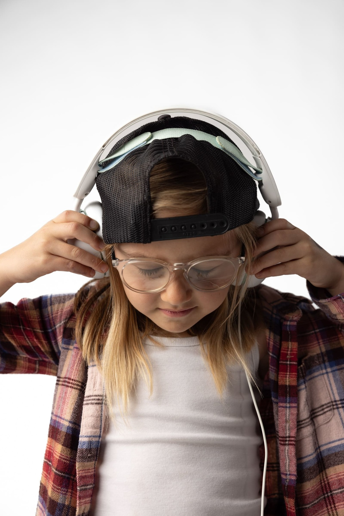 Dziewczyna w okularach i czapce reguluje słuchawki. Ma na sobie koszulę w kratę i biały podkoszulek. Białe tło.
