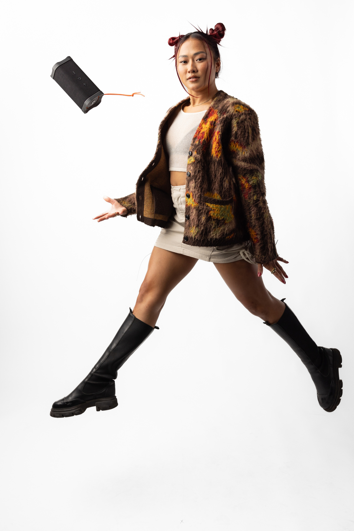 Frau springt mit Lautsprecher. Trägt Jacke, Rock und Stiefel. Weißer Hintergrund.