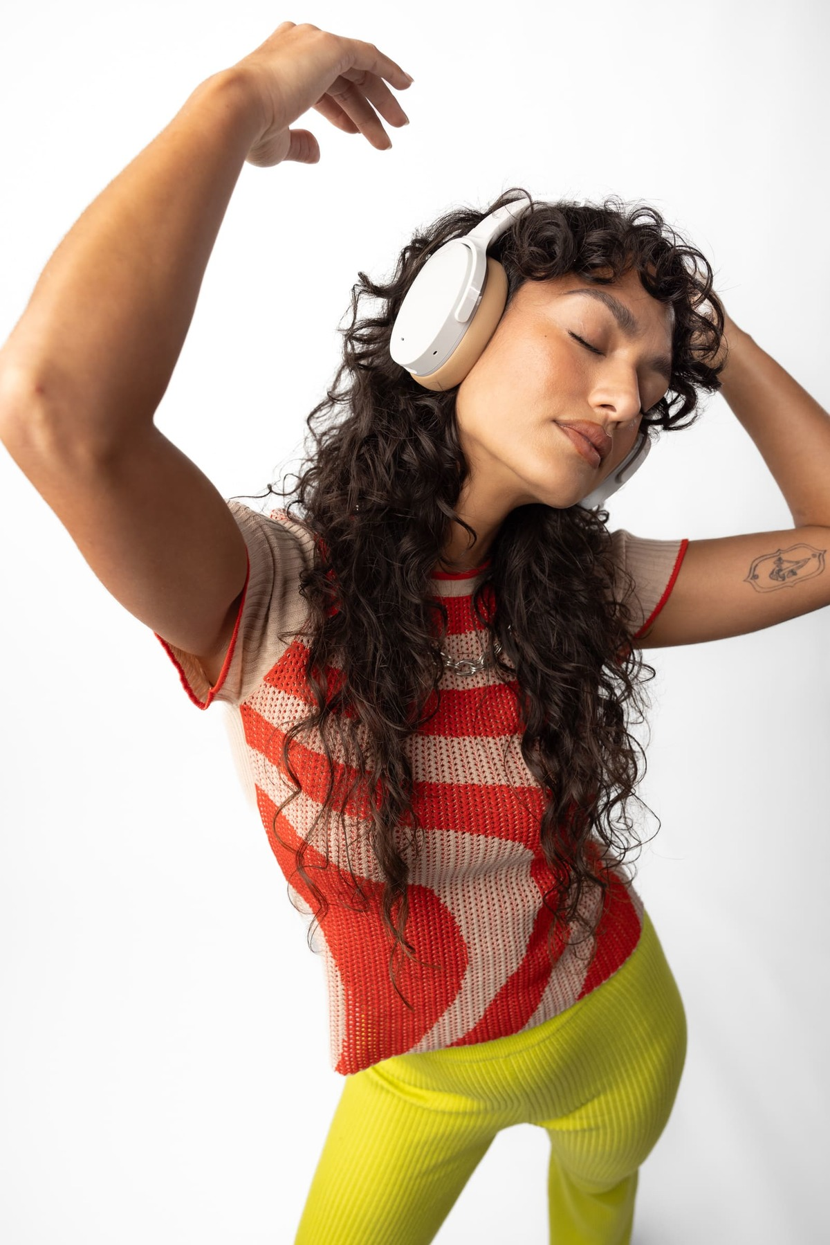 Kobieta z białymi słuchawkami, podniesionymi ramionami, zamkniętymi oczami, w czerwono-beżowej koszuli.