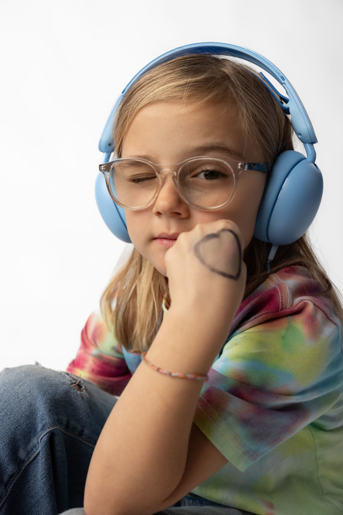 Niña con auriculares azules, gafas y un corazón en la mano. Guiña un ojo.