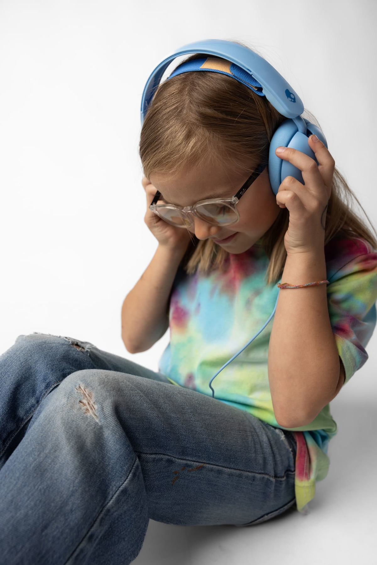 Niña con gafas y auriculares azules. Vaqueros y camiseta teñida. Fondo blanco.