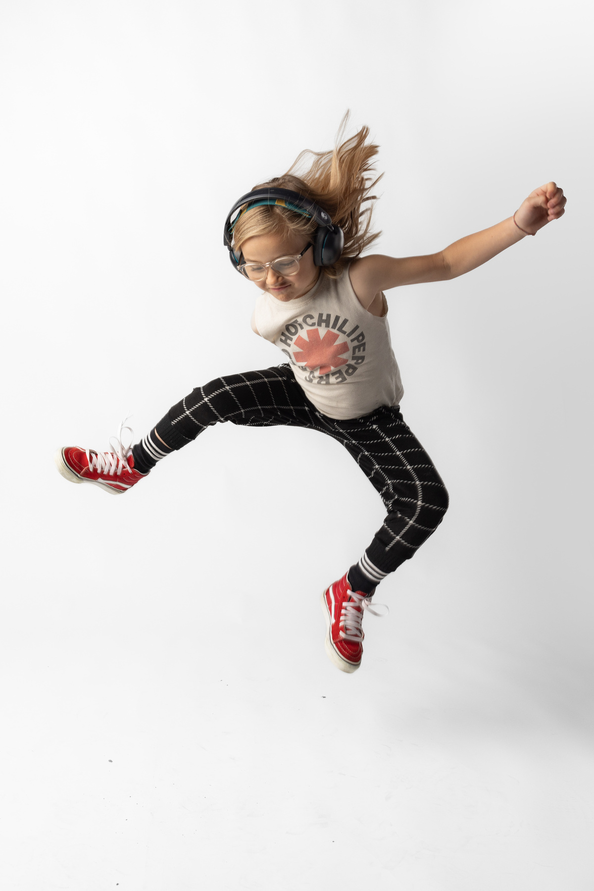 Dziewczyna skacze w słuchawkach, okularach, tank topie i spodniach w kratę. Czerwone buty. Białe tło.