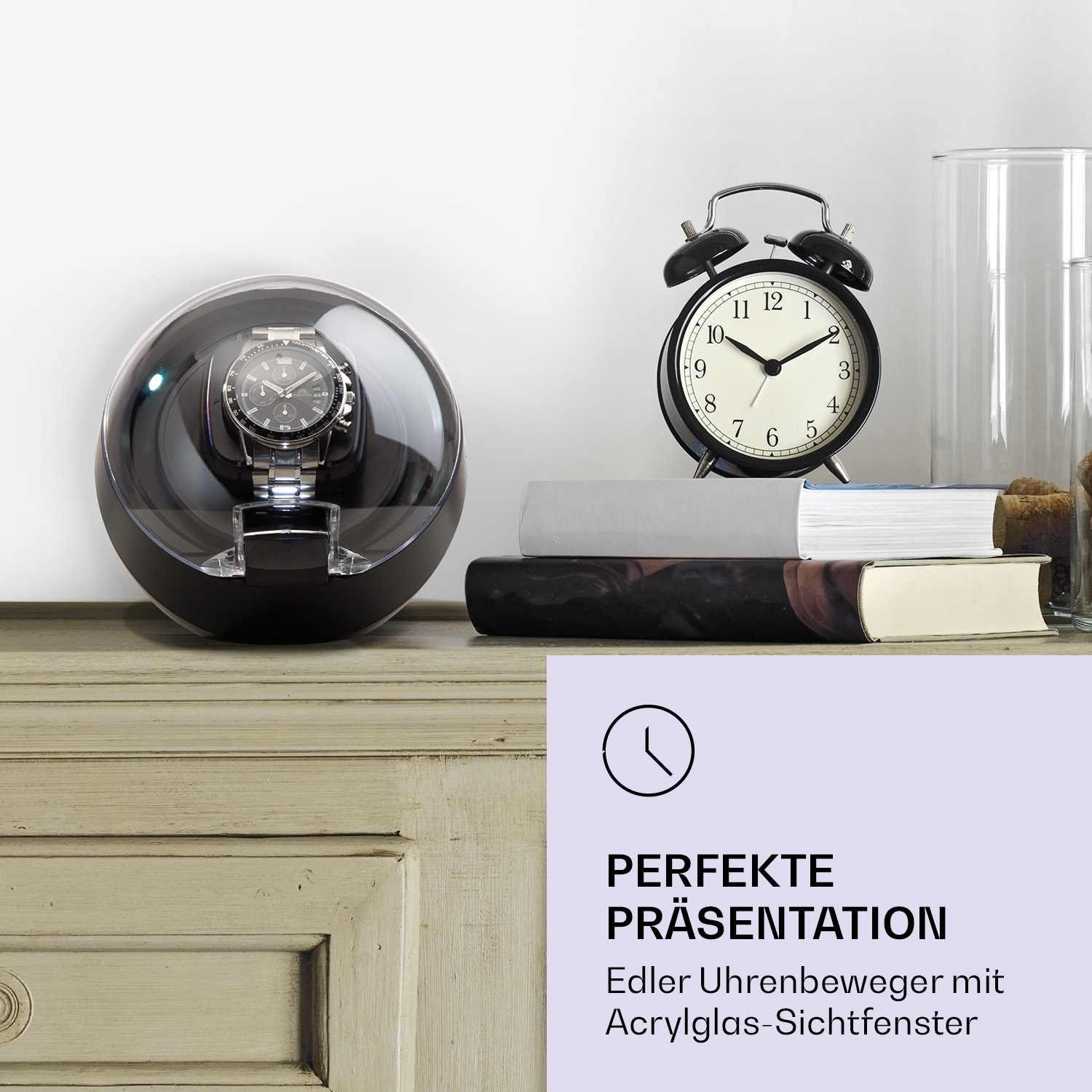 Eine Uhr in einem Glasvitrine auf einem Holztisch. Ein schwarzer Wecker und Bücher sind in der Nähe.
