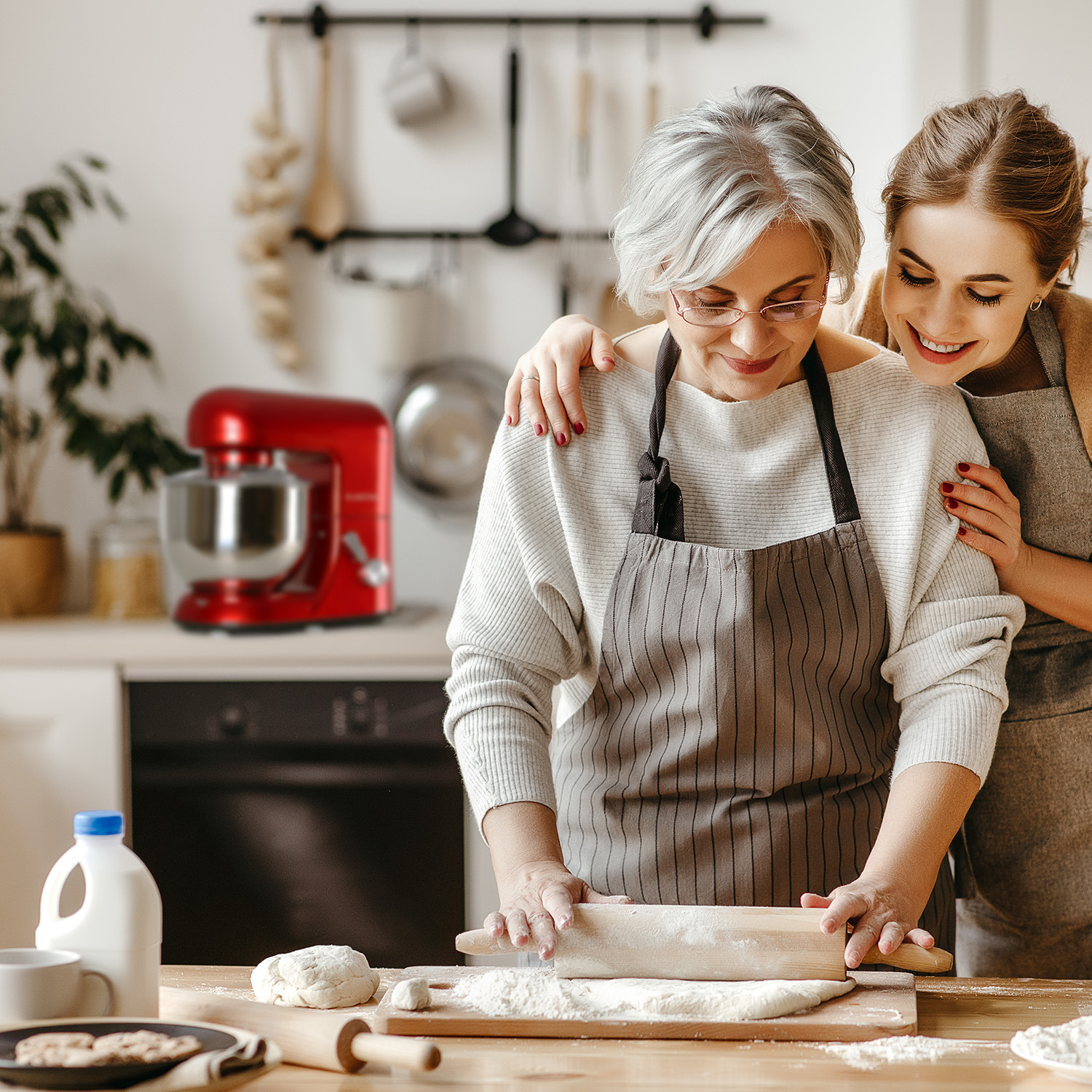Zwei Frauen backen. Eine rollt Teig, die andere umarmt sie. Ein roter Standmixer ist im Hintergrund.
