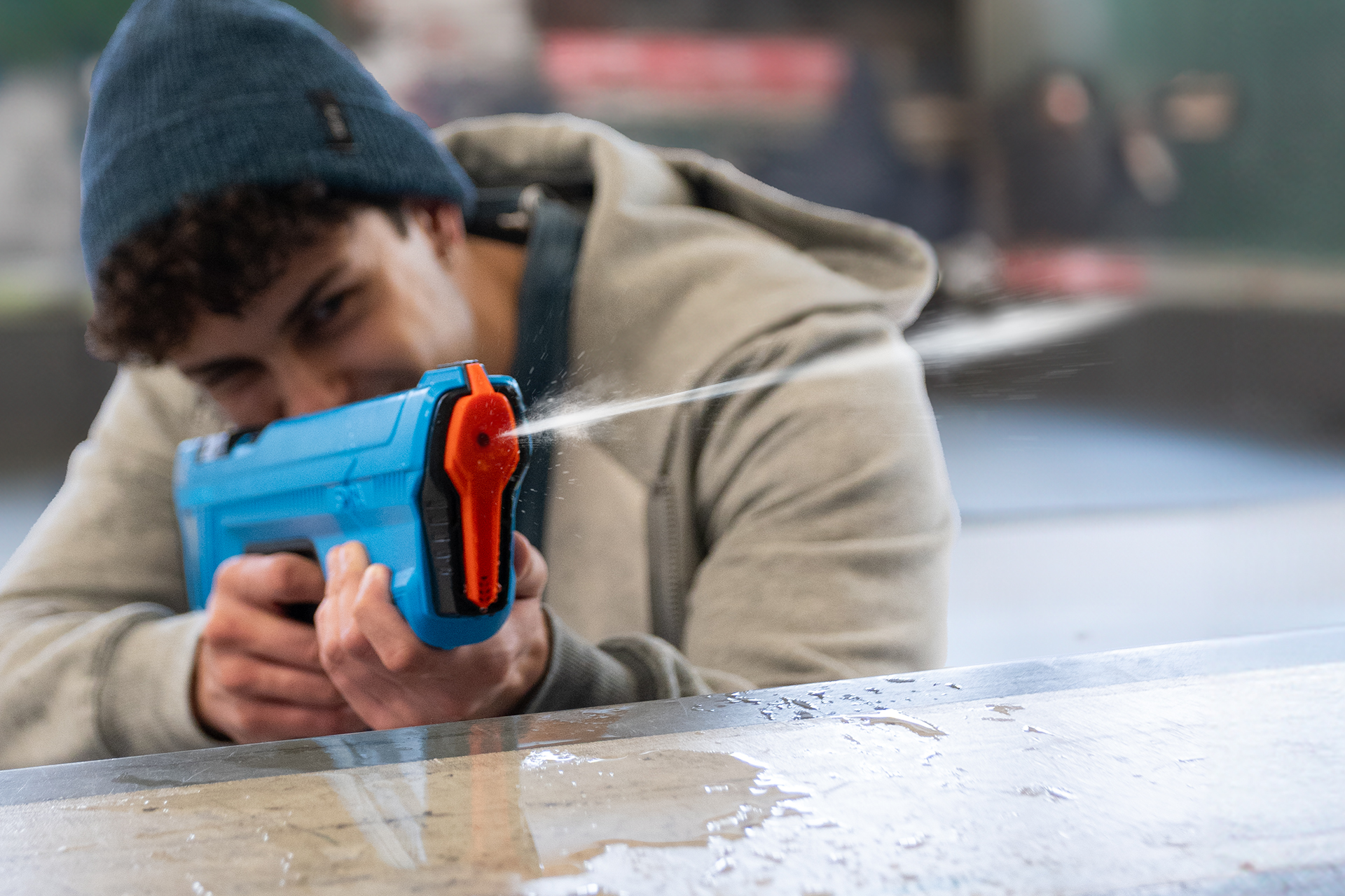 Eine Person zielt mit einer blauen Wasserpistole und sprüht Wasser. Die Person trägt eine blaue Mütze und eine graue Jacke.