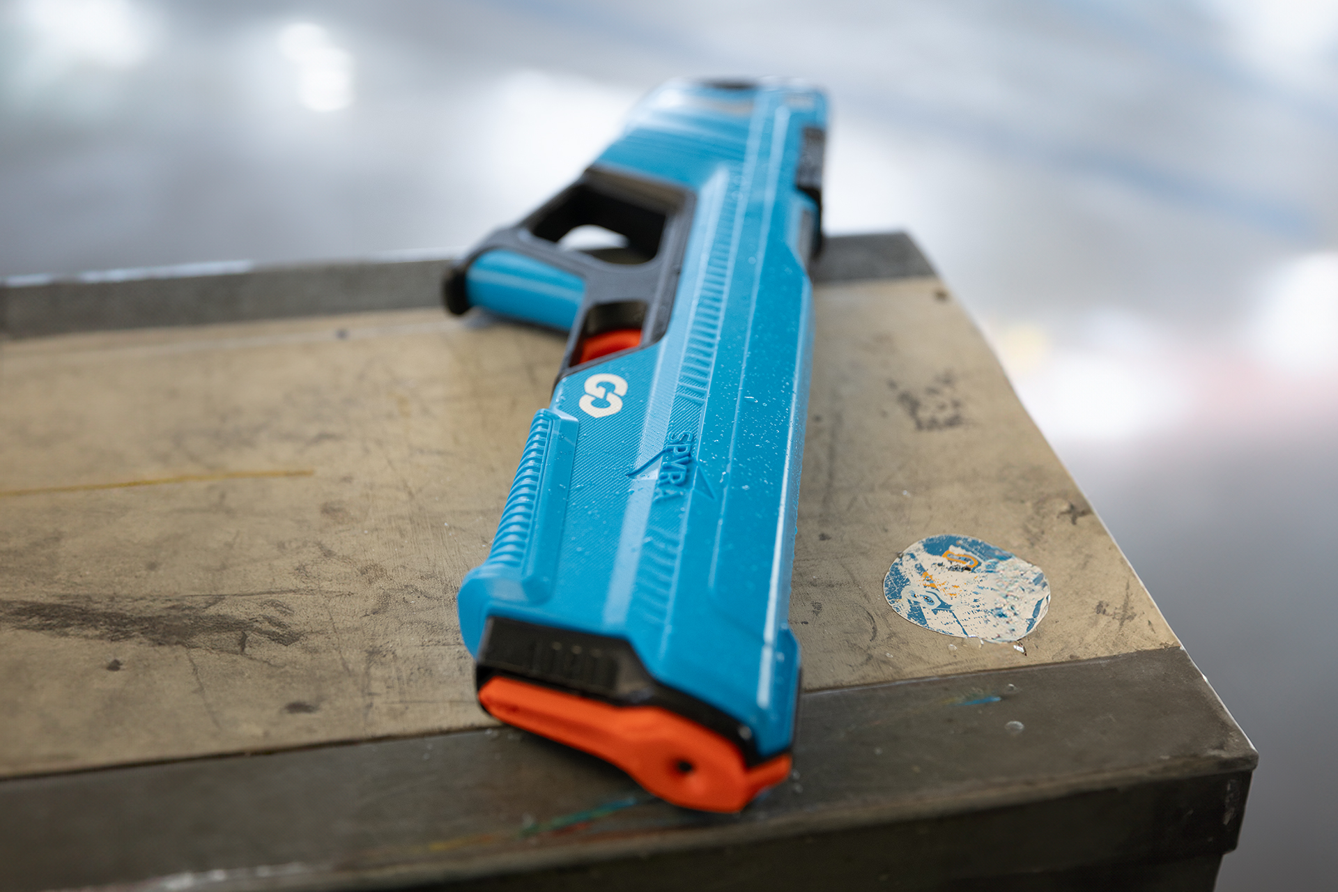 Eine blau-schwarze Wasserpistole liegt auf einer Metalloberfläche. Die Pistole hat eine orangefarbene Düse und einen weißen Aufkleber.