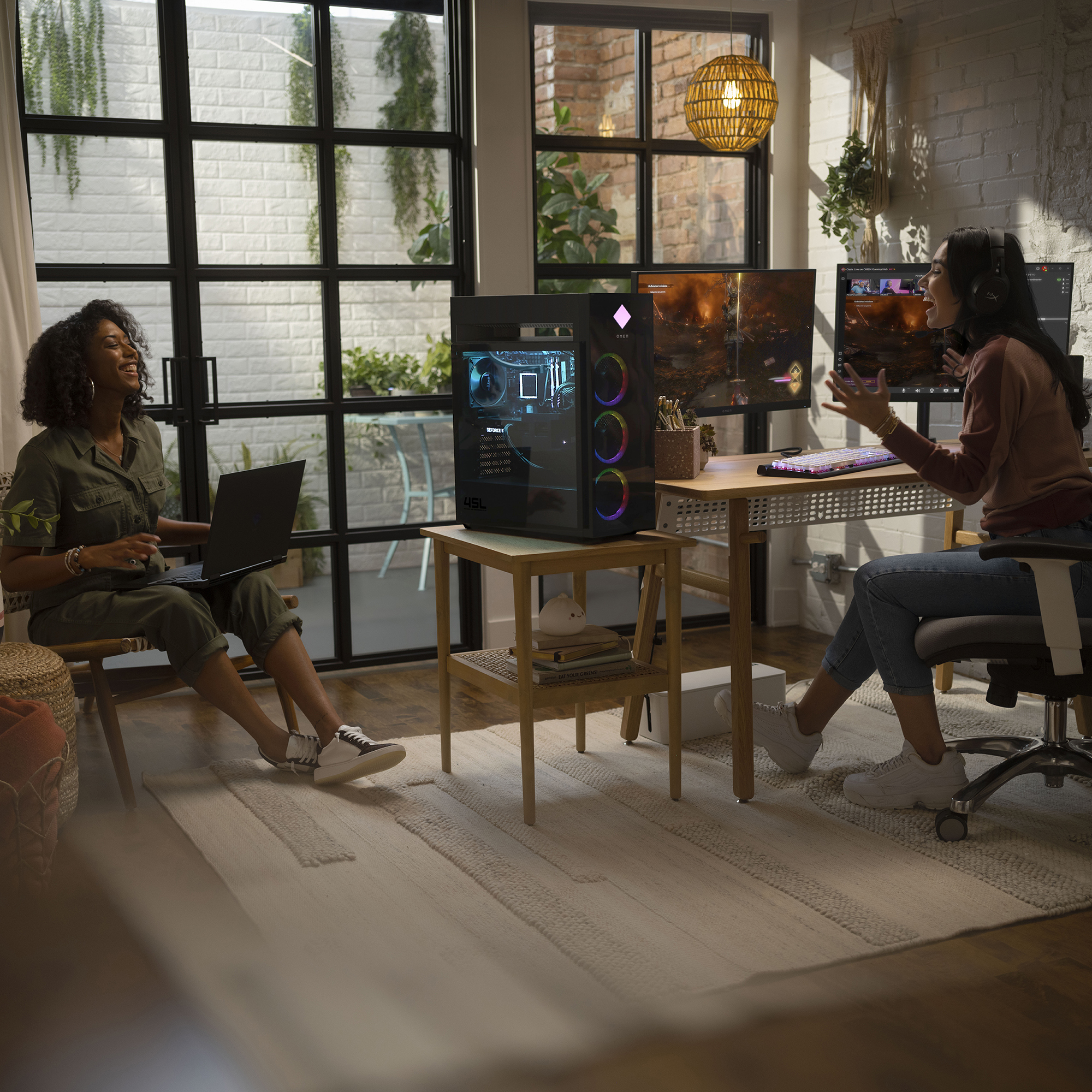 Dwie kobiety w dobrze oświetlonym pokoju, jedna używa laptopa, druga gra na komputerze stacjonarnym z kolorowymi światłami.