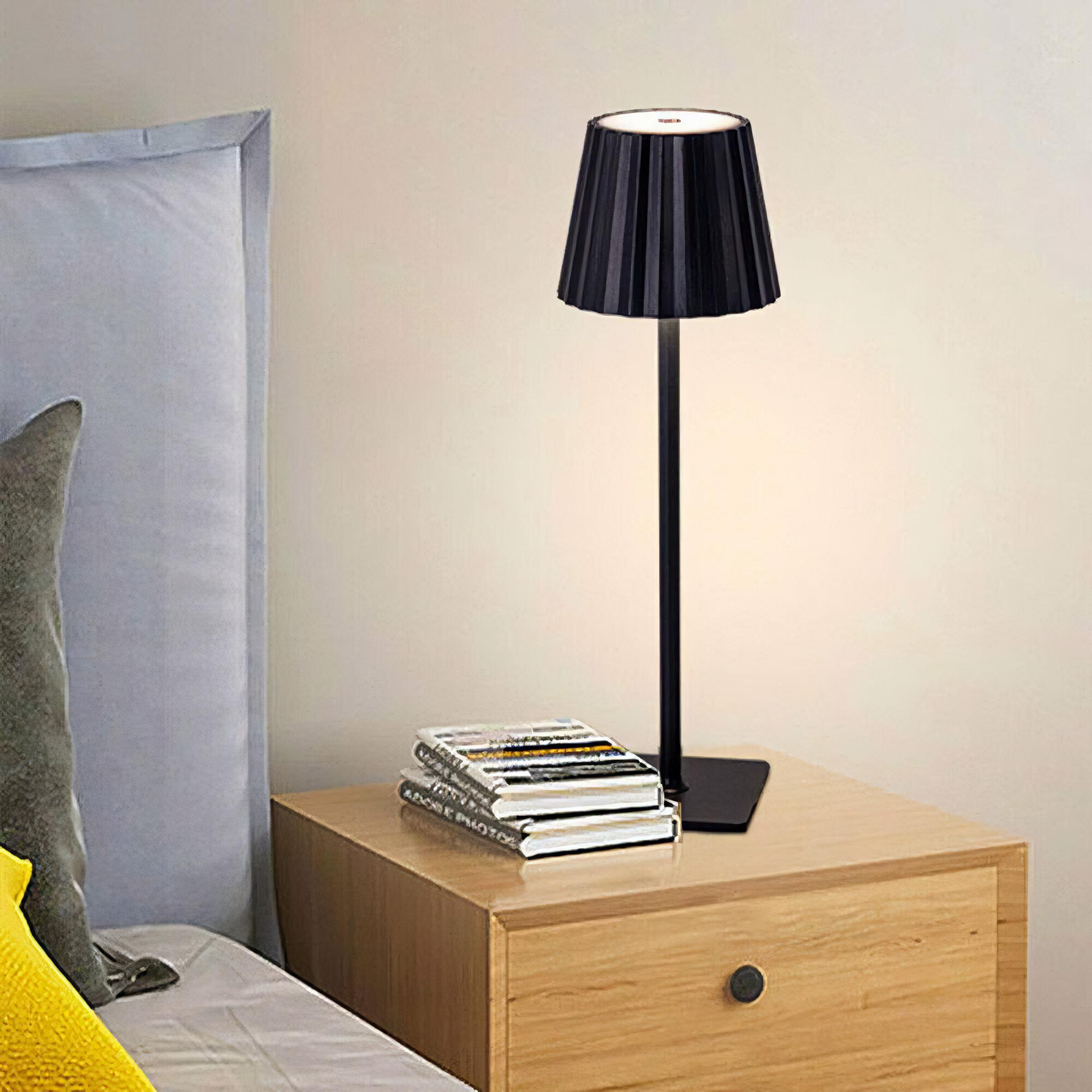Eine schwarze Lampe mit plissiertem Schirm, die auf einem Nachttisch neben Büchern und einem Bett steht.